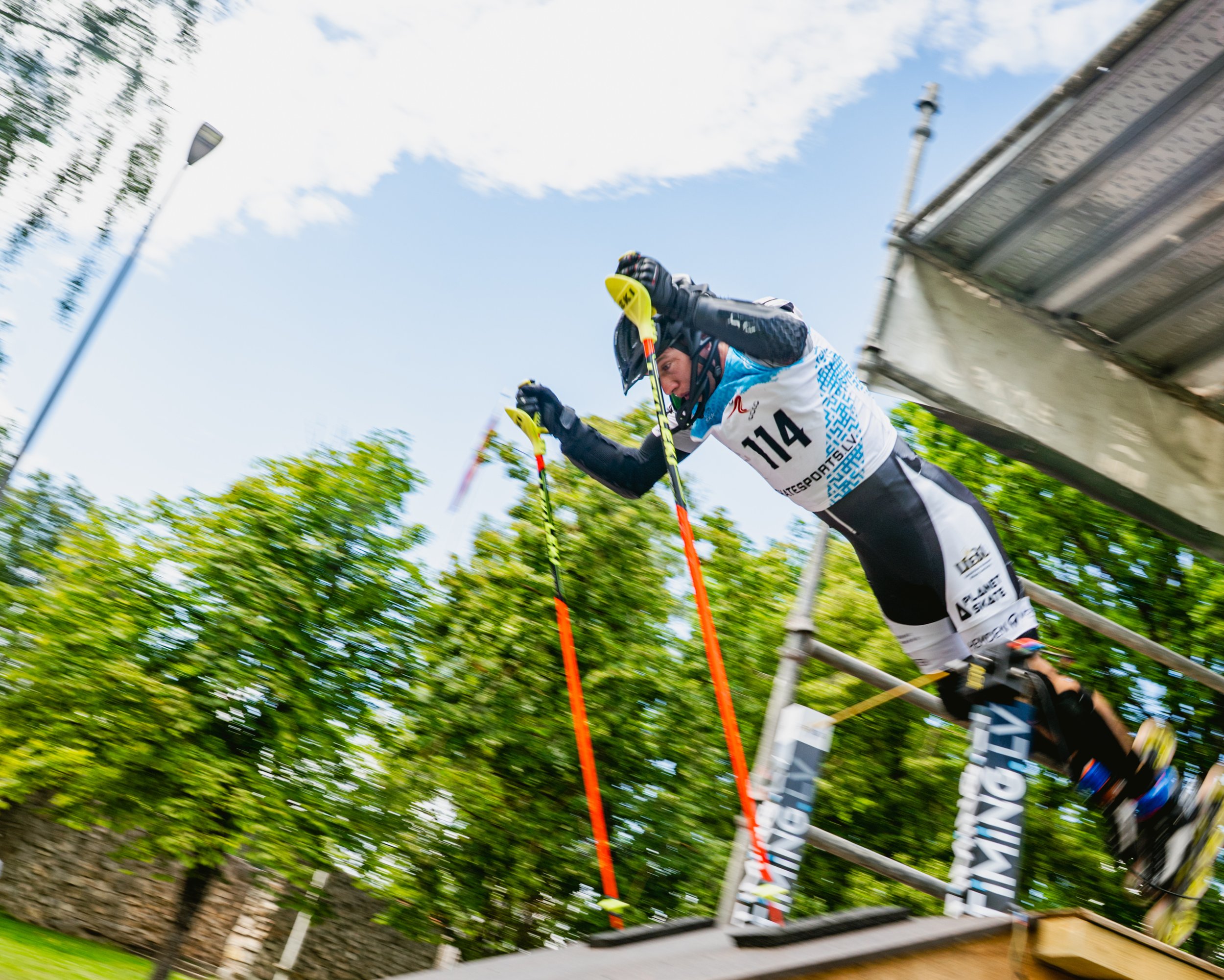 Inline Alpine. A person wearing a helmet and racing gear participating in a sports event, holding ski poles, with the background showing a blue sky, clouds, trees, and a structure.