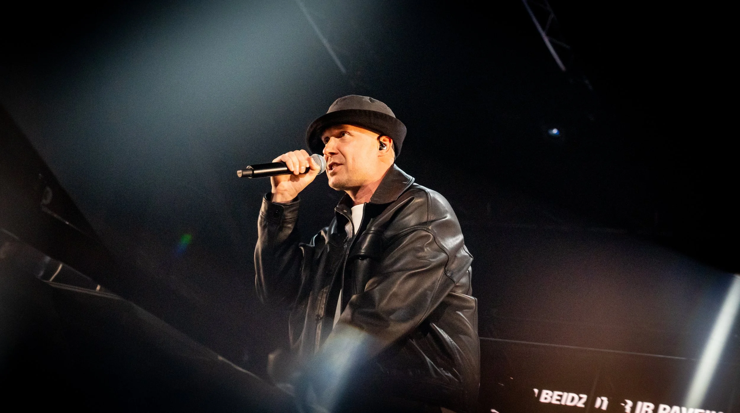 A man wearing a black hat and leather jacket performing on stage, singing into a microphone, with a dark background.