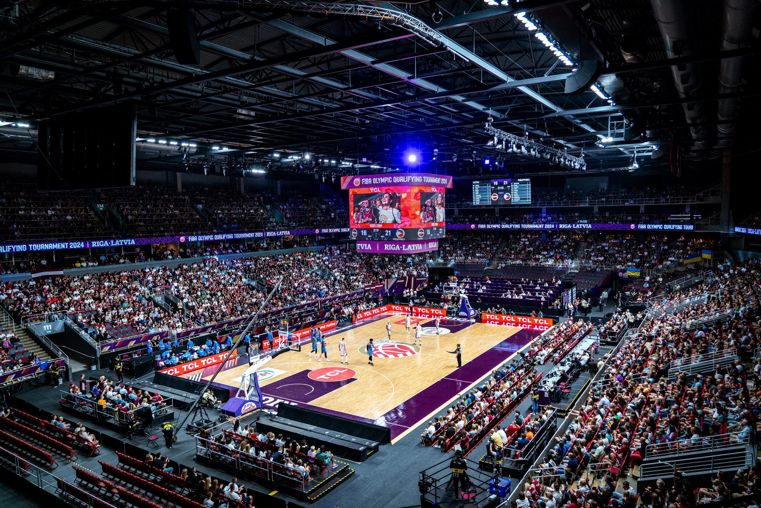Indoor basketball game at the FIBA Olympic Qualifying Tournament 2024, with players on the court, crowds in the stands, and large electronic scoreboards.