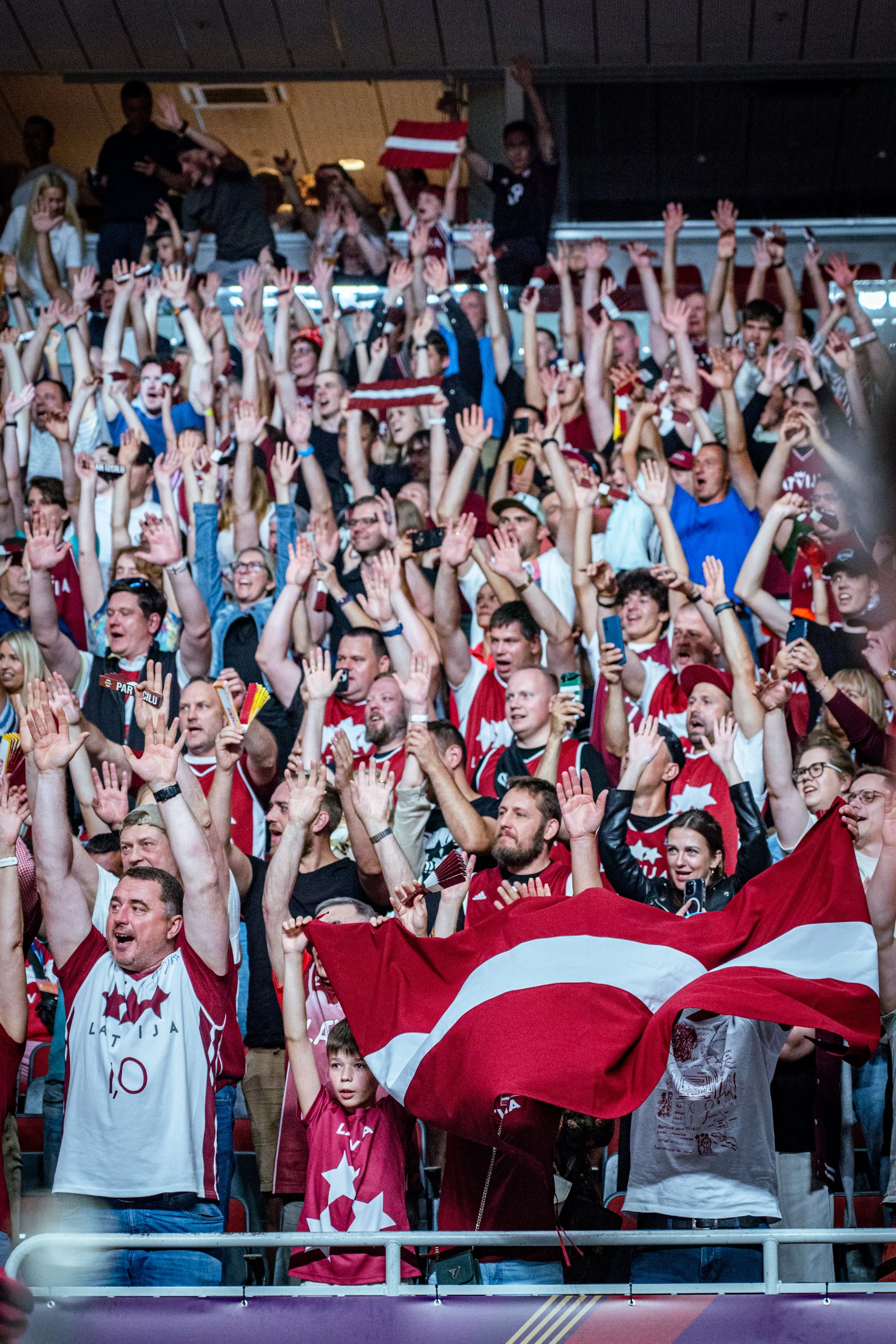 A crowd of enthusiastic sports fans, many wearing red and white jerseys with stars, celebrating in a stadium. Some hold flags, upraise hands, or take photos. A person in the middle holds a red-and-white scarf, and another is waving a large flag. The 