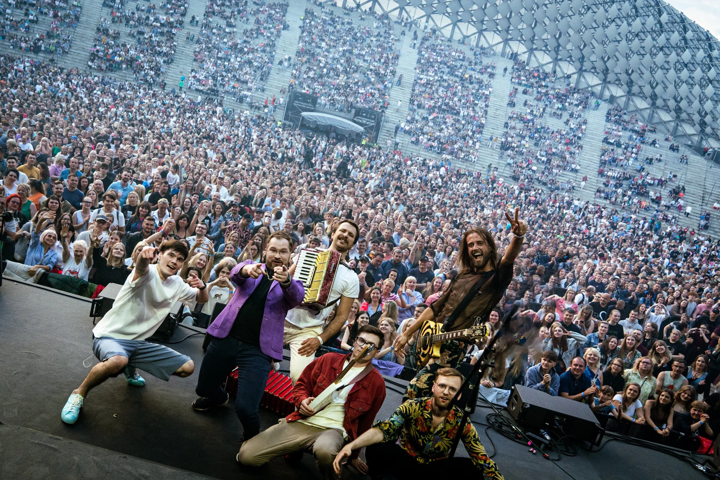 A large crowd of people attending a concert at an outdoor stadium, with performers on stage in the foreground, including a man with a guitar, a man with a keyboard, and a man with an accordion, all smiling and engaging with the audience.