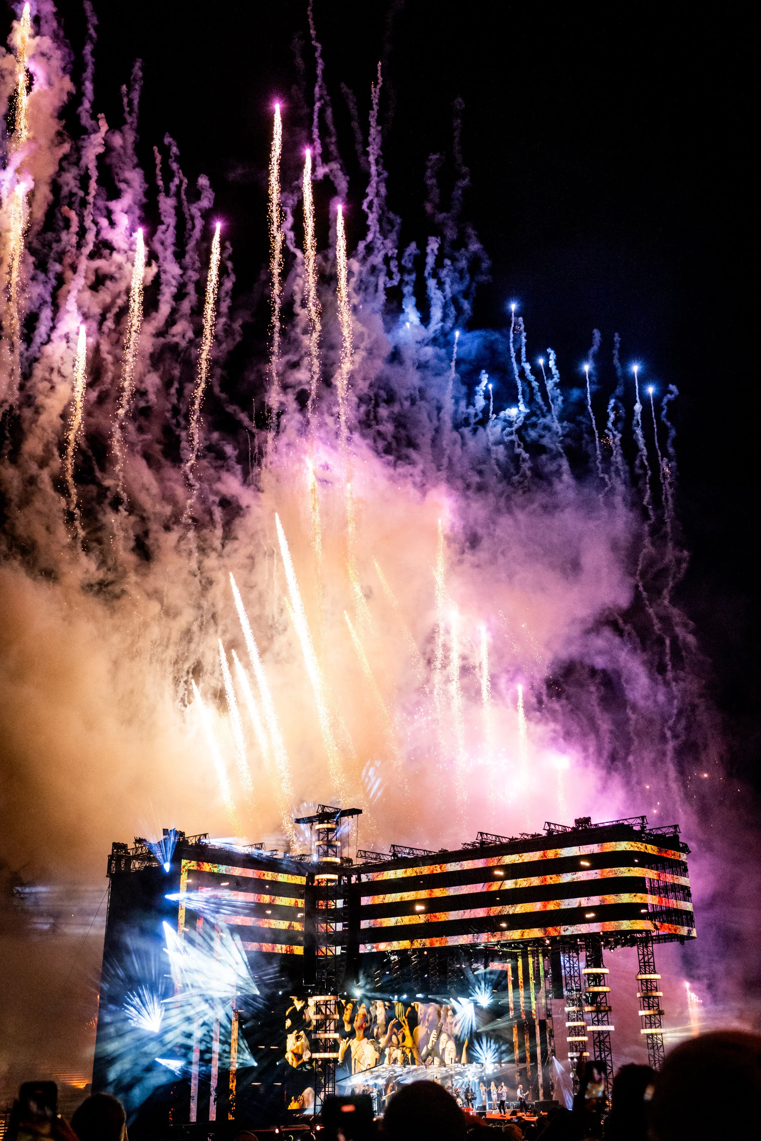 Nighttime fireworks display lights up the sky over a concert stage with large screen showing performers, illuminated by colorful stage lighting and smoke.