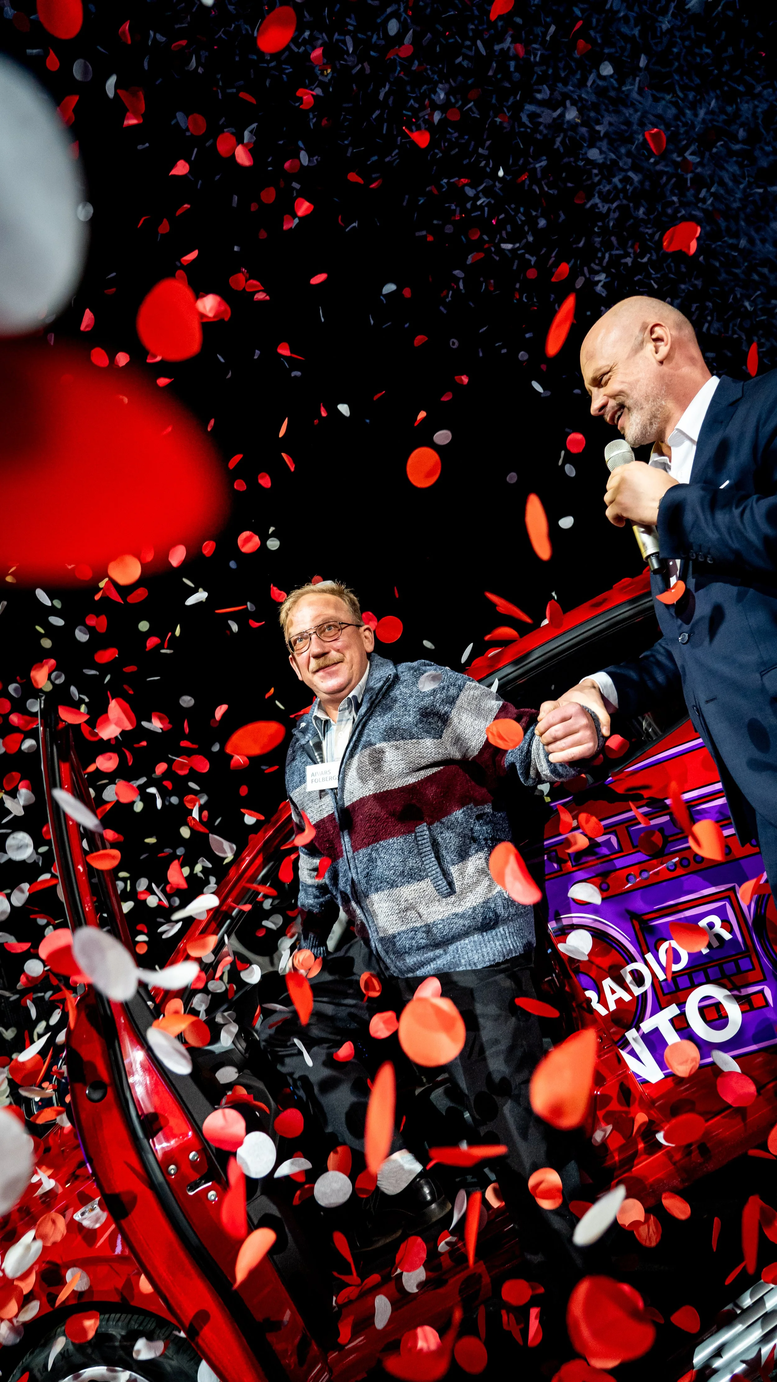 Two men standing beside a red vehicle with an open door, celebrating with red and white confetti falling around them. One man is wearing glasses and a striped sweater, the other is dressed in a suit and holding a microphone.