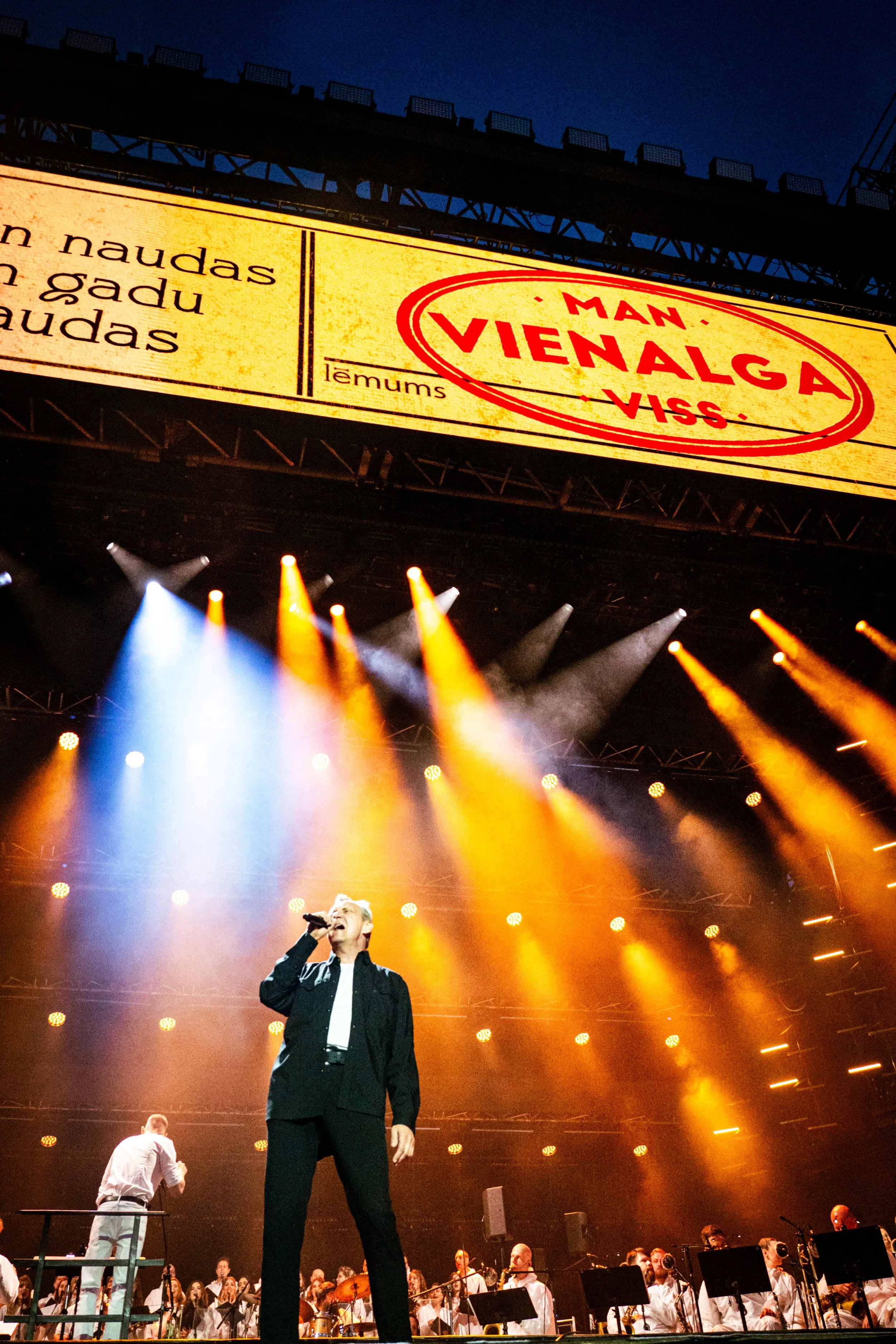 A singer performing on stage with an orchestra, colorful lights, and a large screen displaying text behind him.