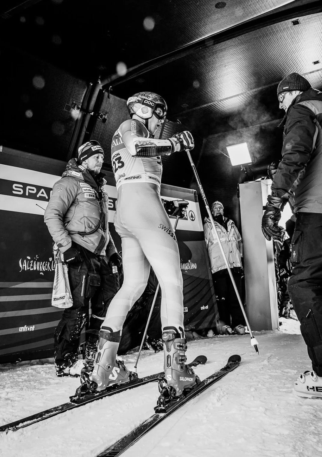 A skier in racing gear with a helmet and goggles standing on skis in a ski lodge, surrounded by people in winter clothing.