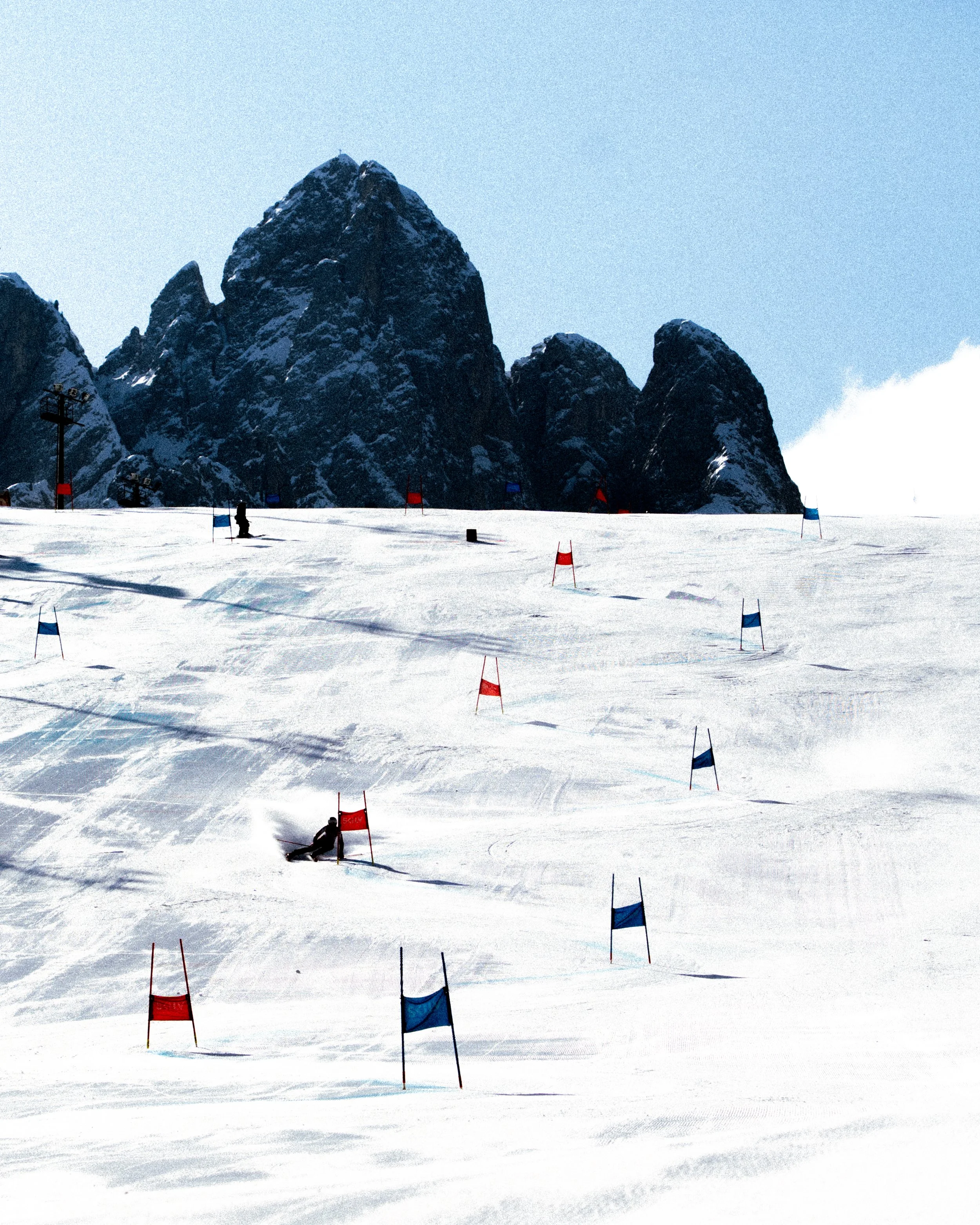 A skier making a turn around a red slalom ski gate on a snow-covered mountain slope with a rocky mountain in the background under a clear blue sky.