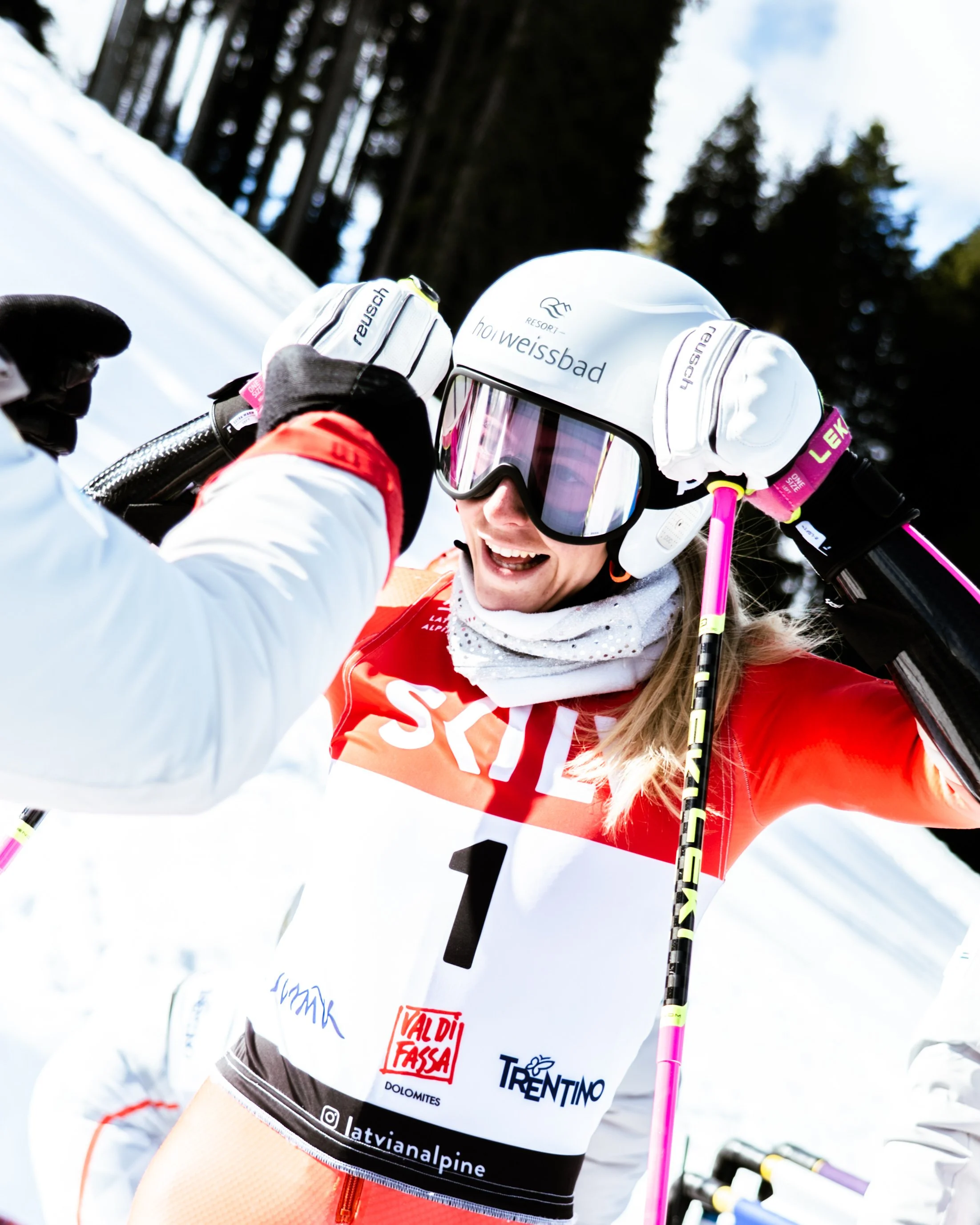 A smiling female skier in a red and white racing suit, holding ski poles, wearing a white helmet with the words "Resort Holweissbad," and sunglasses, with snowy mountain landscape and trees in the background.