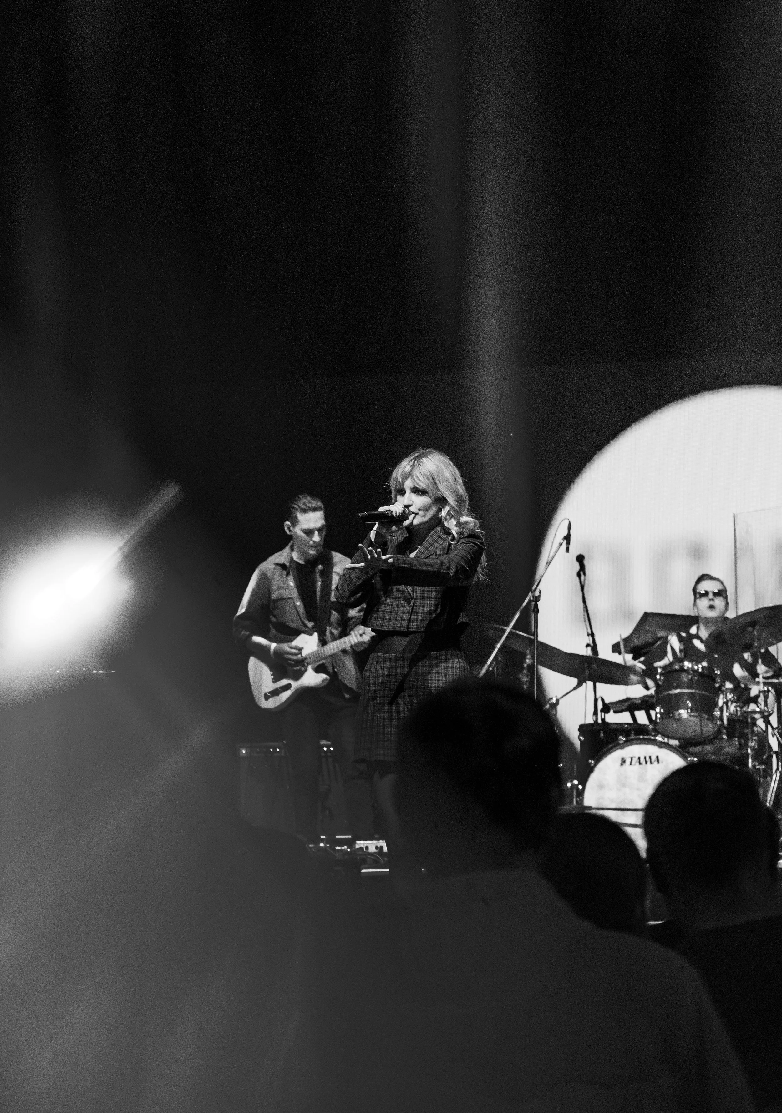 Black and white photo of a woman singing into a microphone, with a guitarist and drummer on stage, performing under stage lights.