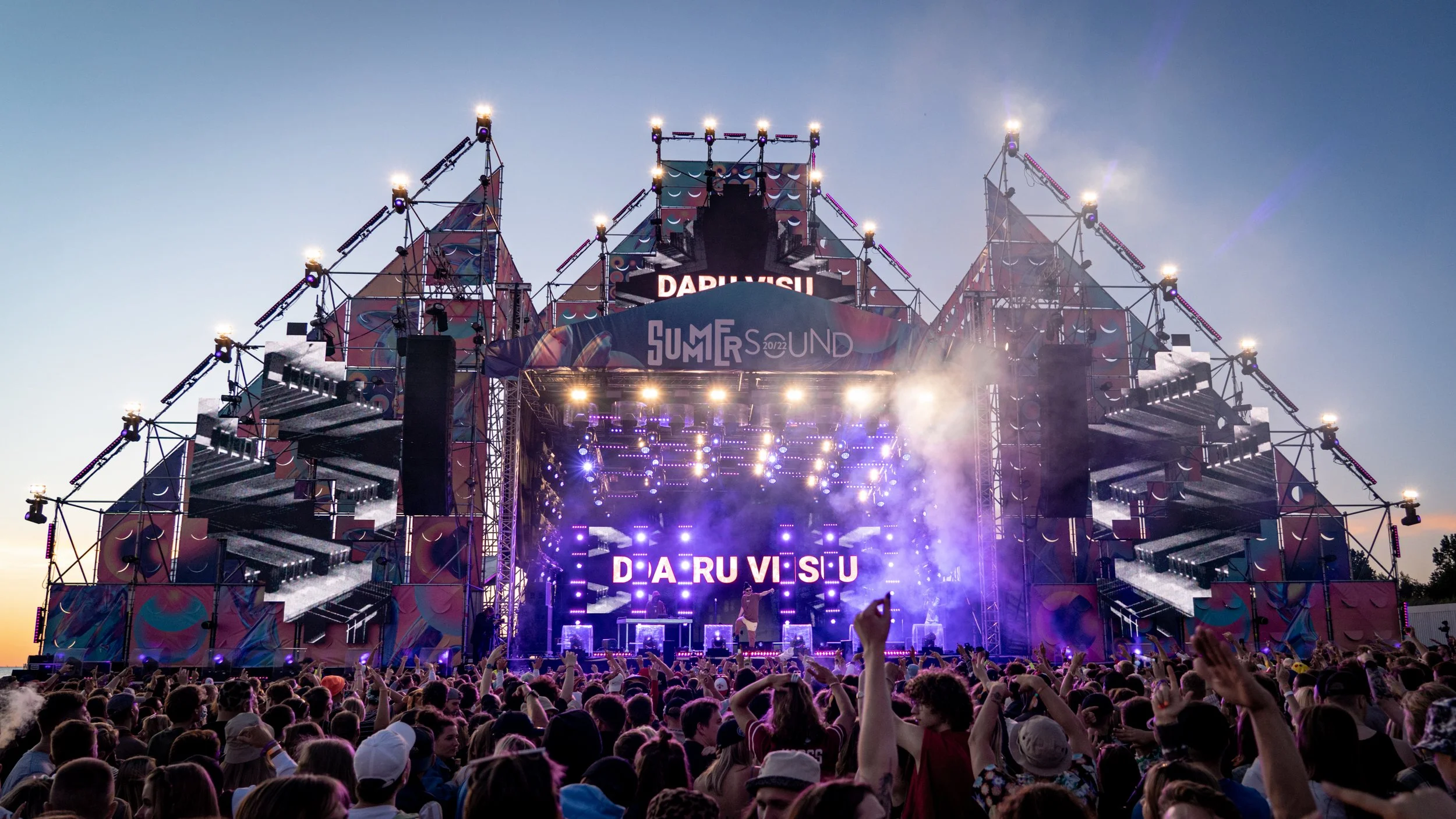 A large outdoor music festival stage with bright lights and a crowd of people dancing and enjoying the concert at sunset.