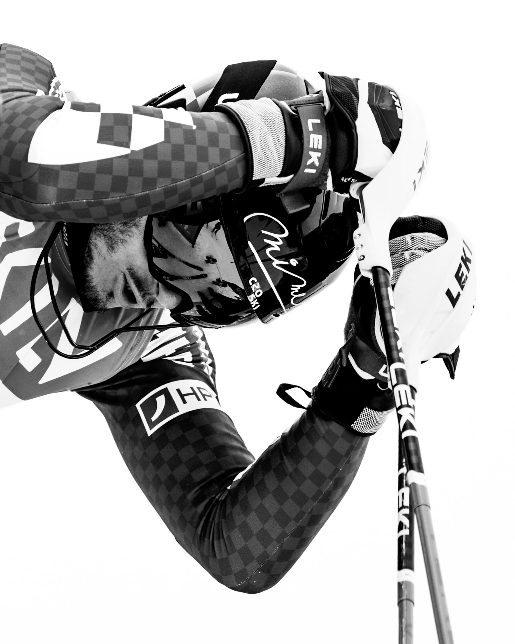 A skier in a checkered jacket and helmet, holding ski poles, appears to be resting or looking down, captured in black and white.