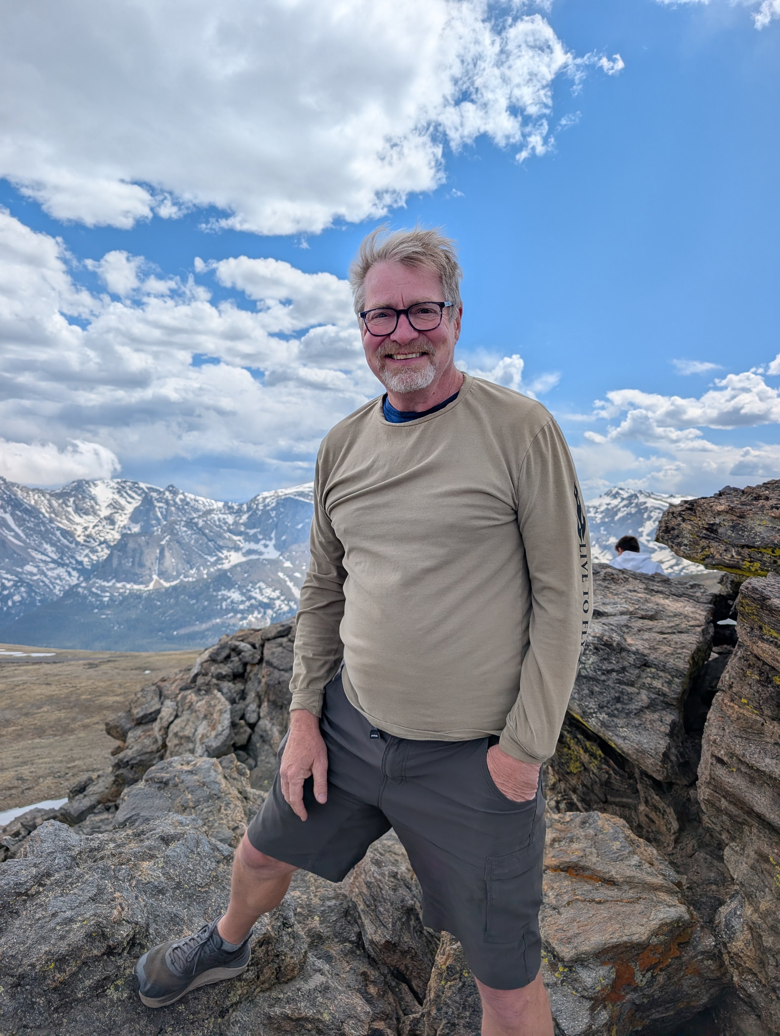 Man wearing glasses standing on rocky terrain with snow-capped mountains and a partly cloudy blue sky behind him