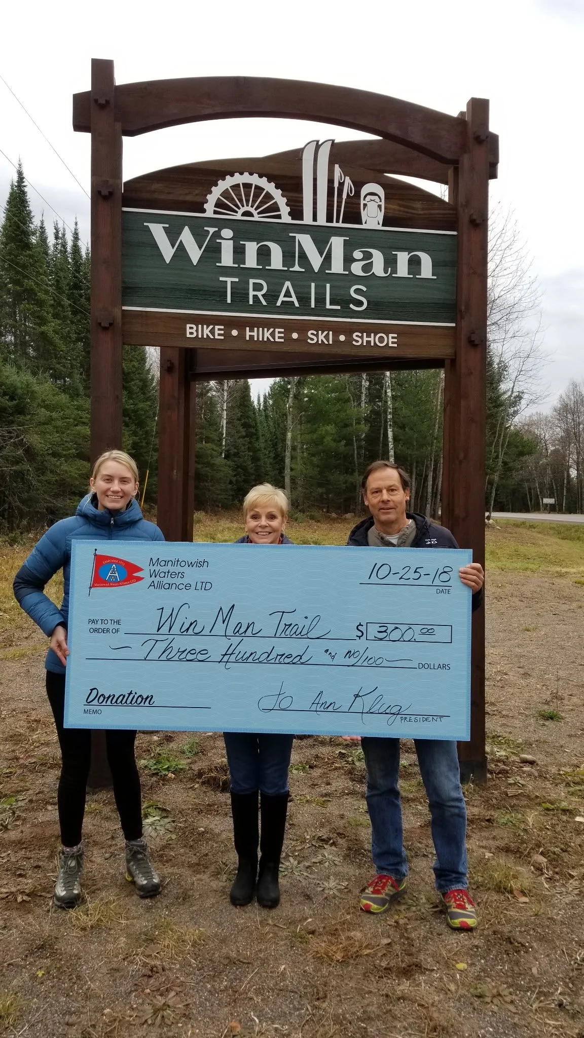 Three people hold a $300 donation check beside the WinMan Trails sign in a wooded Wisconsin setting.