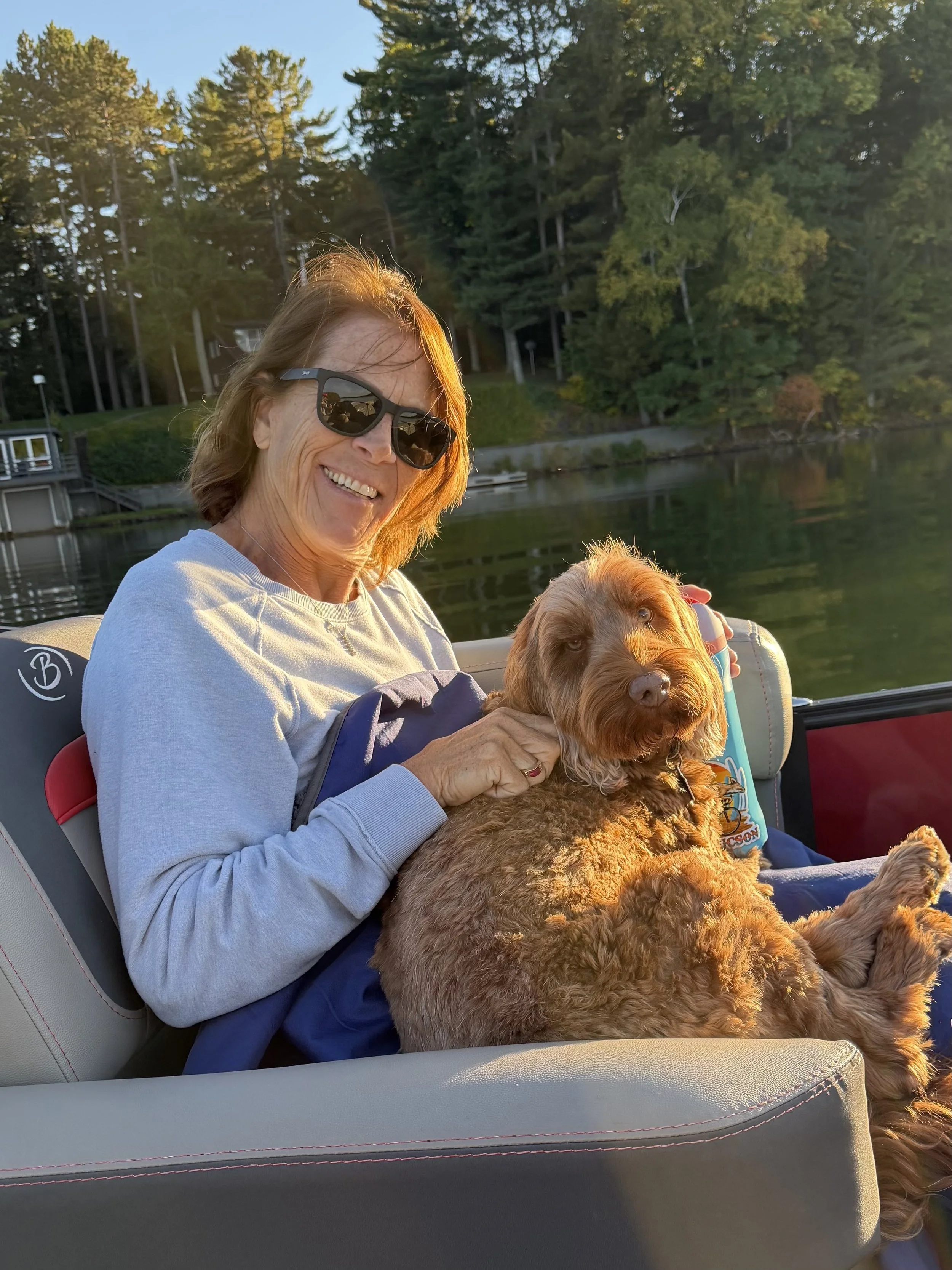 Smiling woman wearing sunglasses holds a curly brown dog on a boat near a tree-lined lake at sunset.