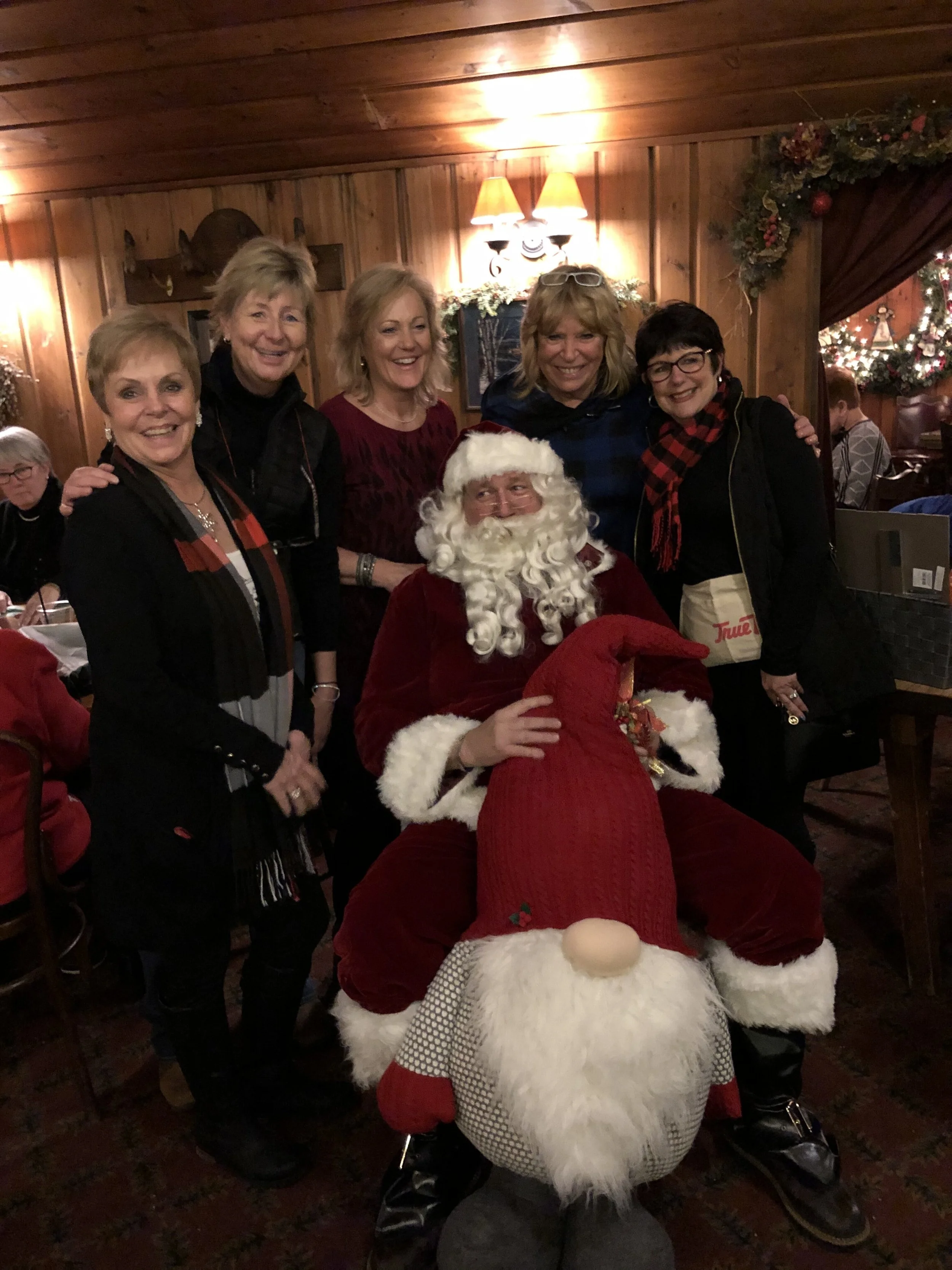 Group of women posing with Santa Claus in a warmly lit, wood-paneled room decorated for Christmas