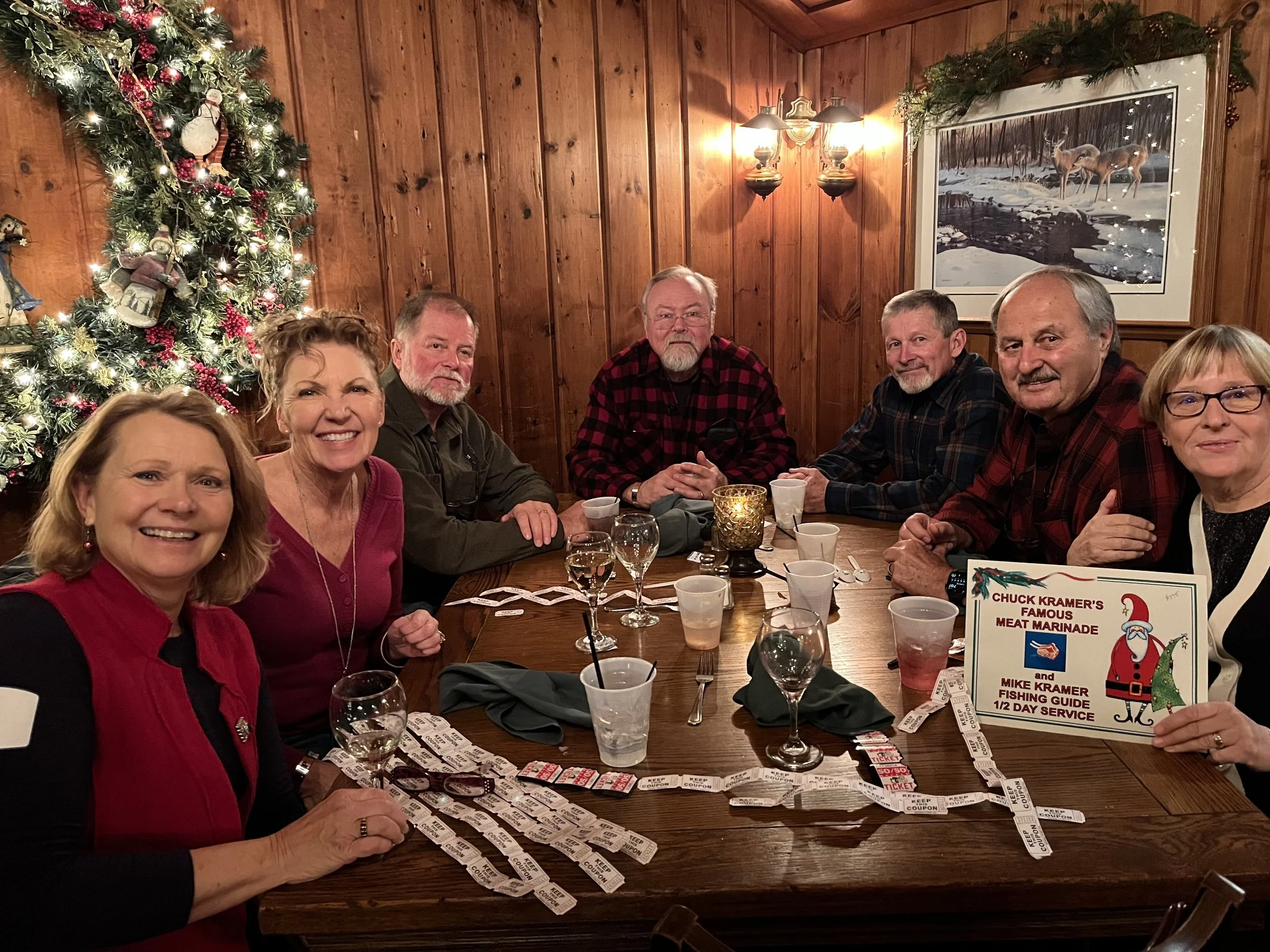 Group of friends celebrating around a restaurant table beside a lit Christmas tree, with holiday decorations, raffle tickets, and a fishing guide sign.