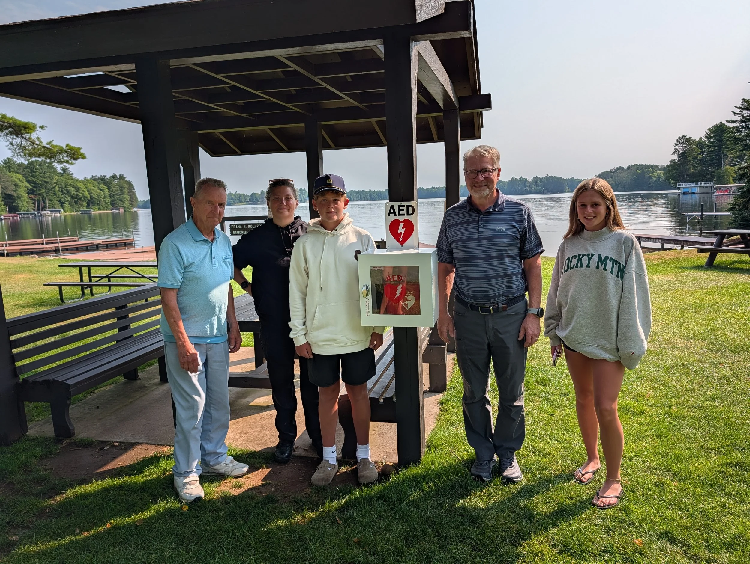 Five people pose beneath a lakeside pavilion beside an AED defibrillator cabinet, with picnic tables and a wooded lake in the background.