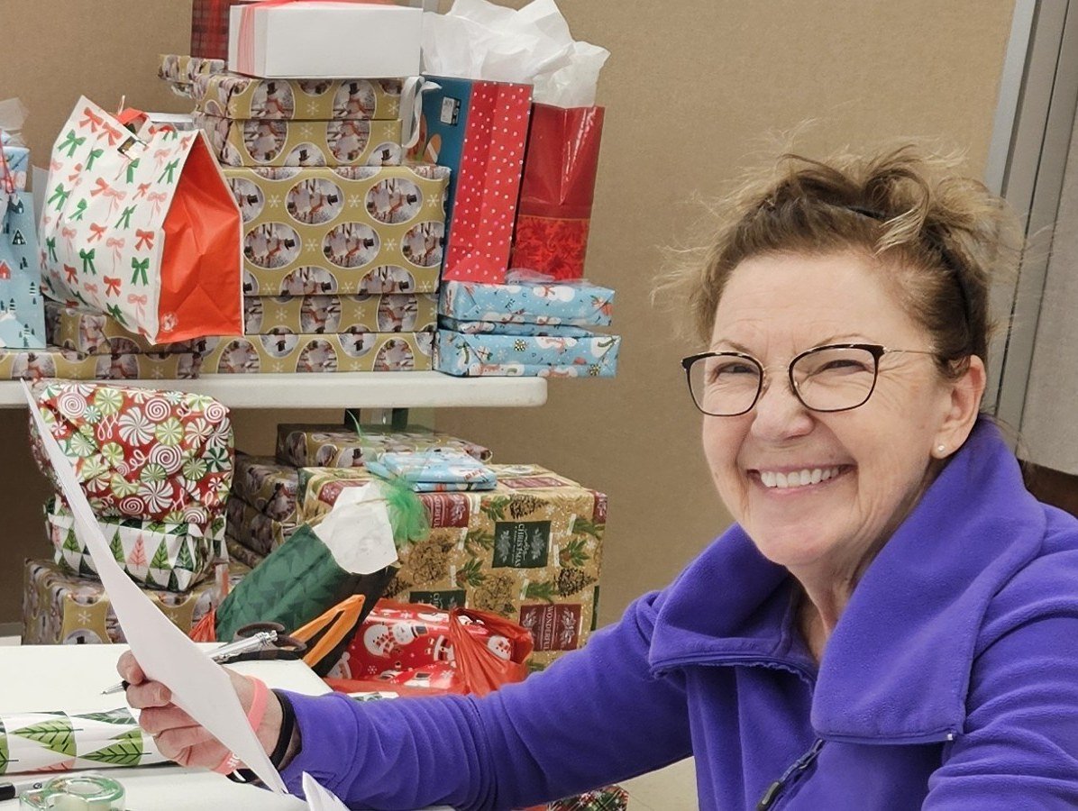 Smiling woman wearing glasses and a purple fleece organizes wrapped Christmas gifts while holding a sheet of paper.