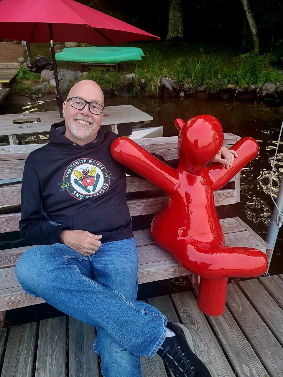 Smiling bald man wearing glasses sits beside a glossy red human-shaped sculpture on a wooden dock by a pond under a red umbrella.
