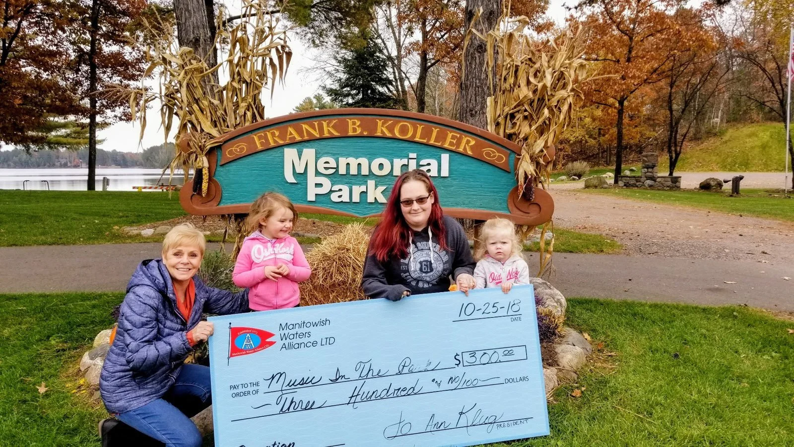 Four people pose with a large $300 ceremonial check in front of the Frank B. Koller Memorial Park sign beside a lake in autumn.
