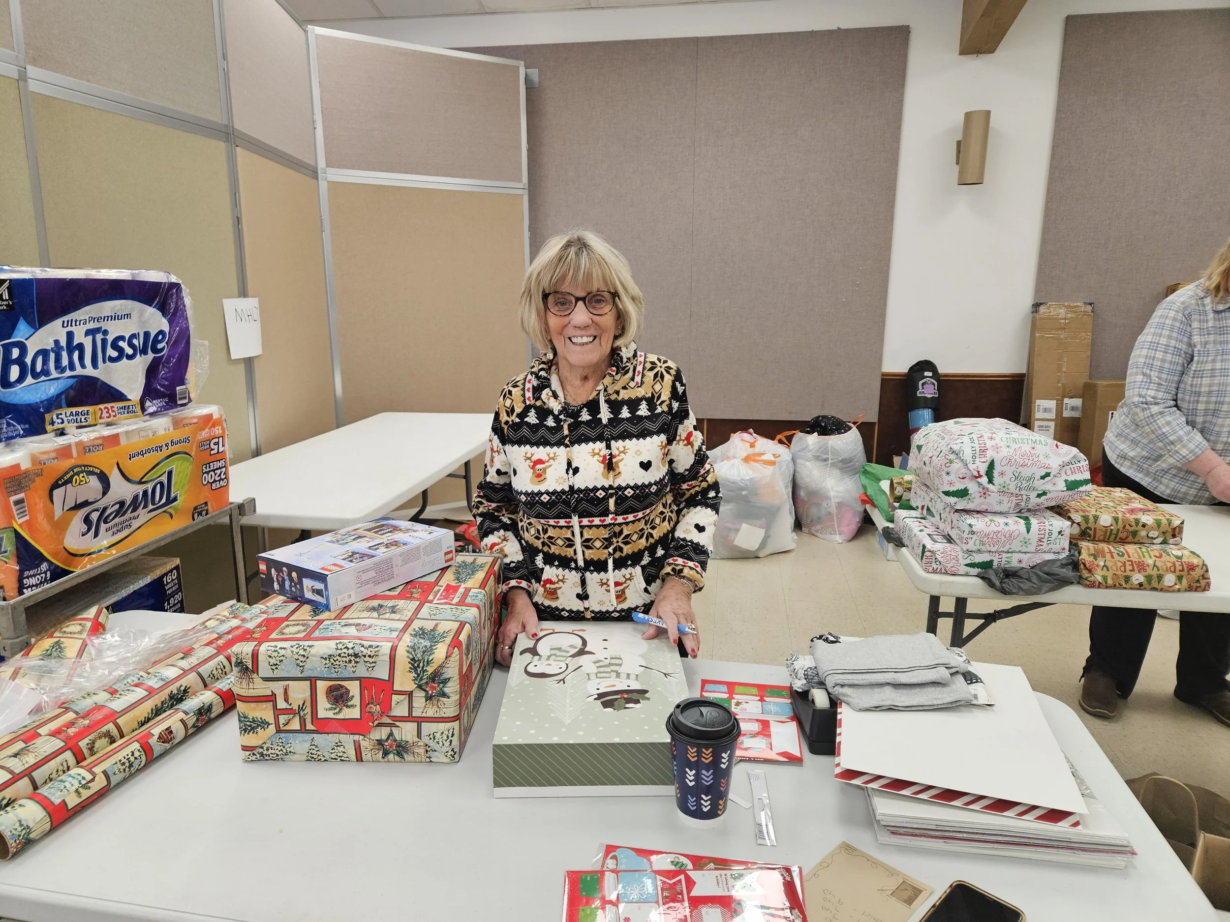 Smiling older woman in a festive Christmas sweater surrounded by wrapped gifts and holiday supplies at a community event.