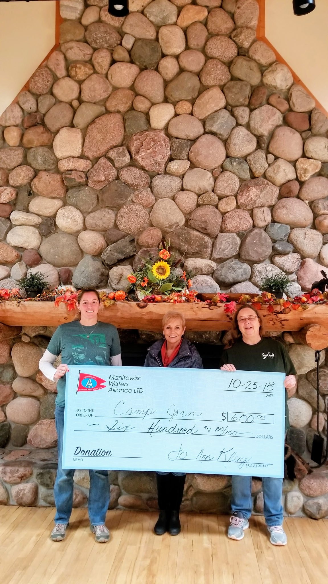 Three women present a $600 donation check from Manitowish Waters Alliance LTD to Camp Jorn in front of a stone fireplace.