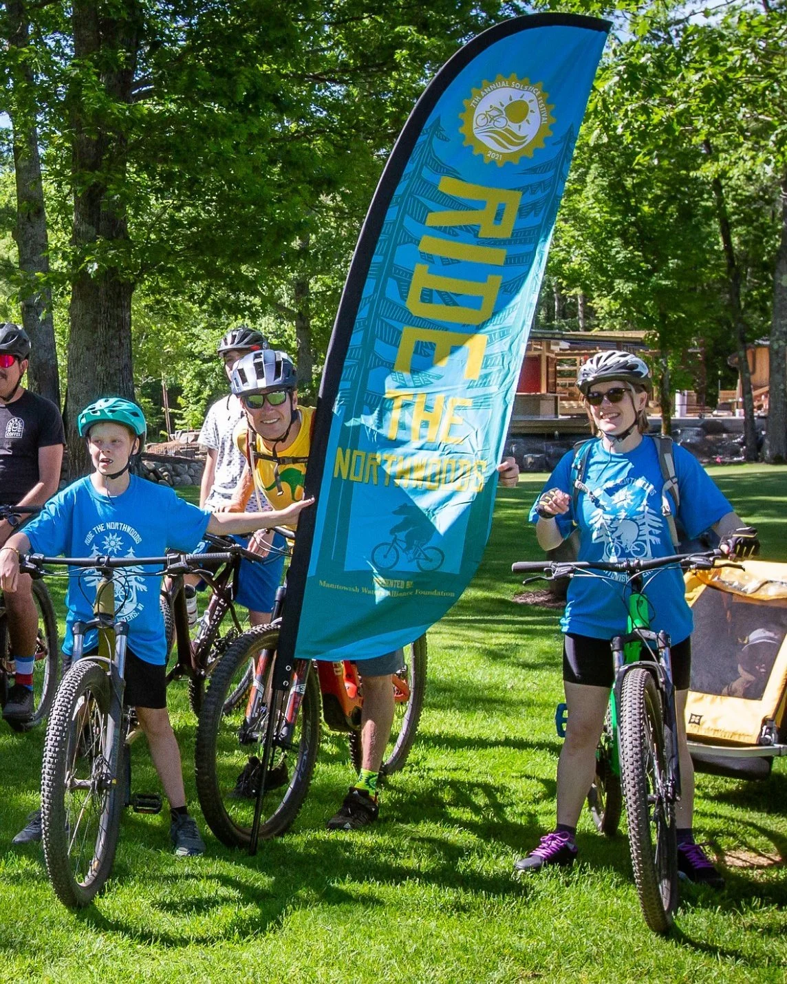 Mountain bikers pose with bicycles beside a blue event flag at an outdoor cycling festival in a wooded park.