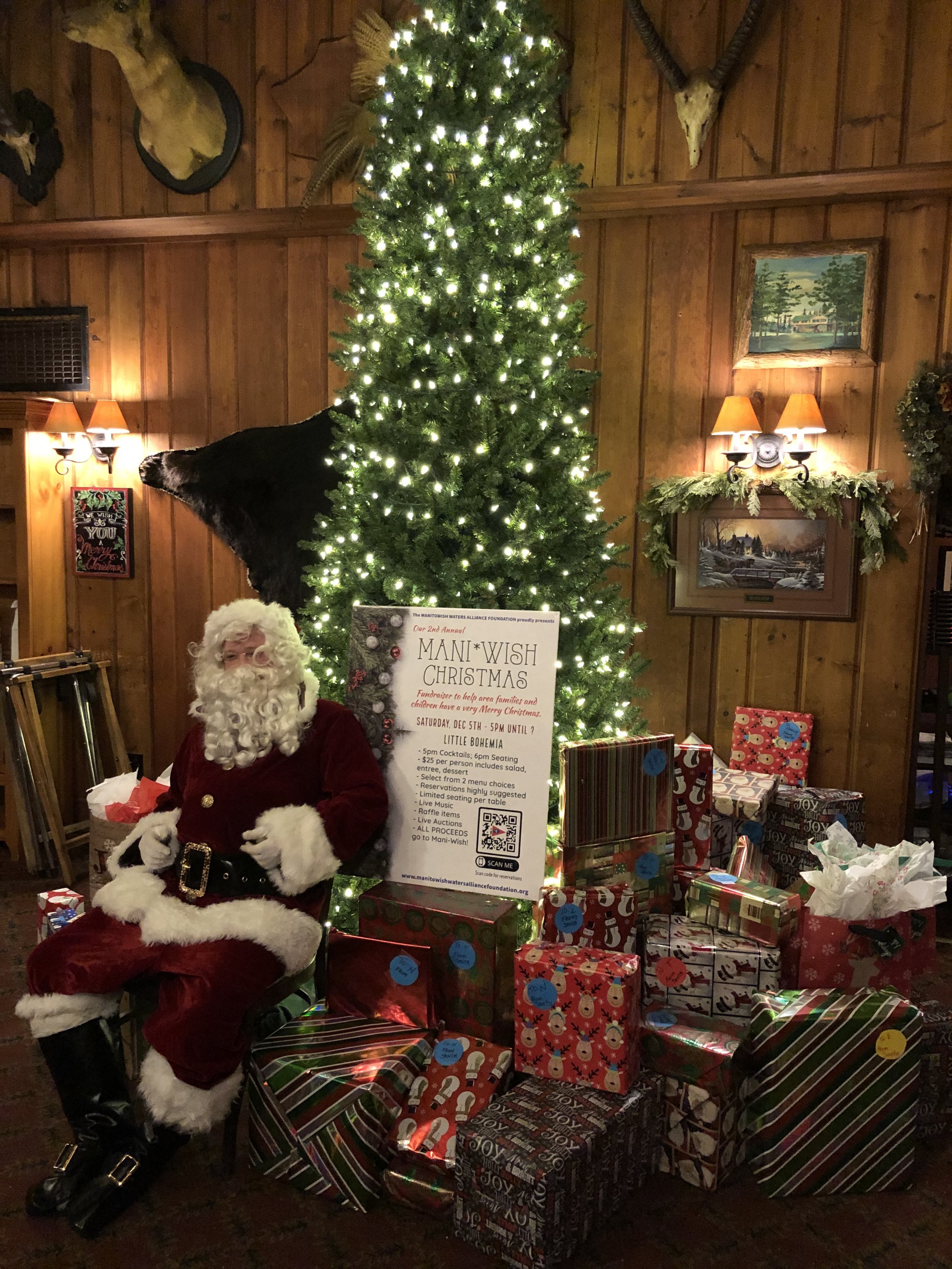 Santa Claus seated beside a lit Christmas tree surrounded by wrapped presents in a rustic wood-paneled lodge.