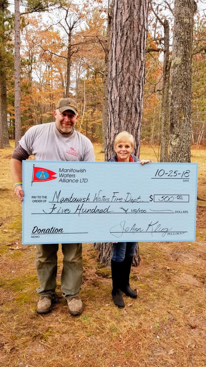 Two people in an autumn forest hold an oversized $500 donation check for the Manitowish Waters Fire Department from Manitowish Waters Alliance LTD.