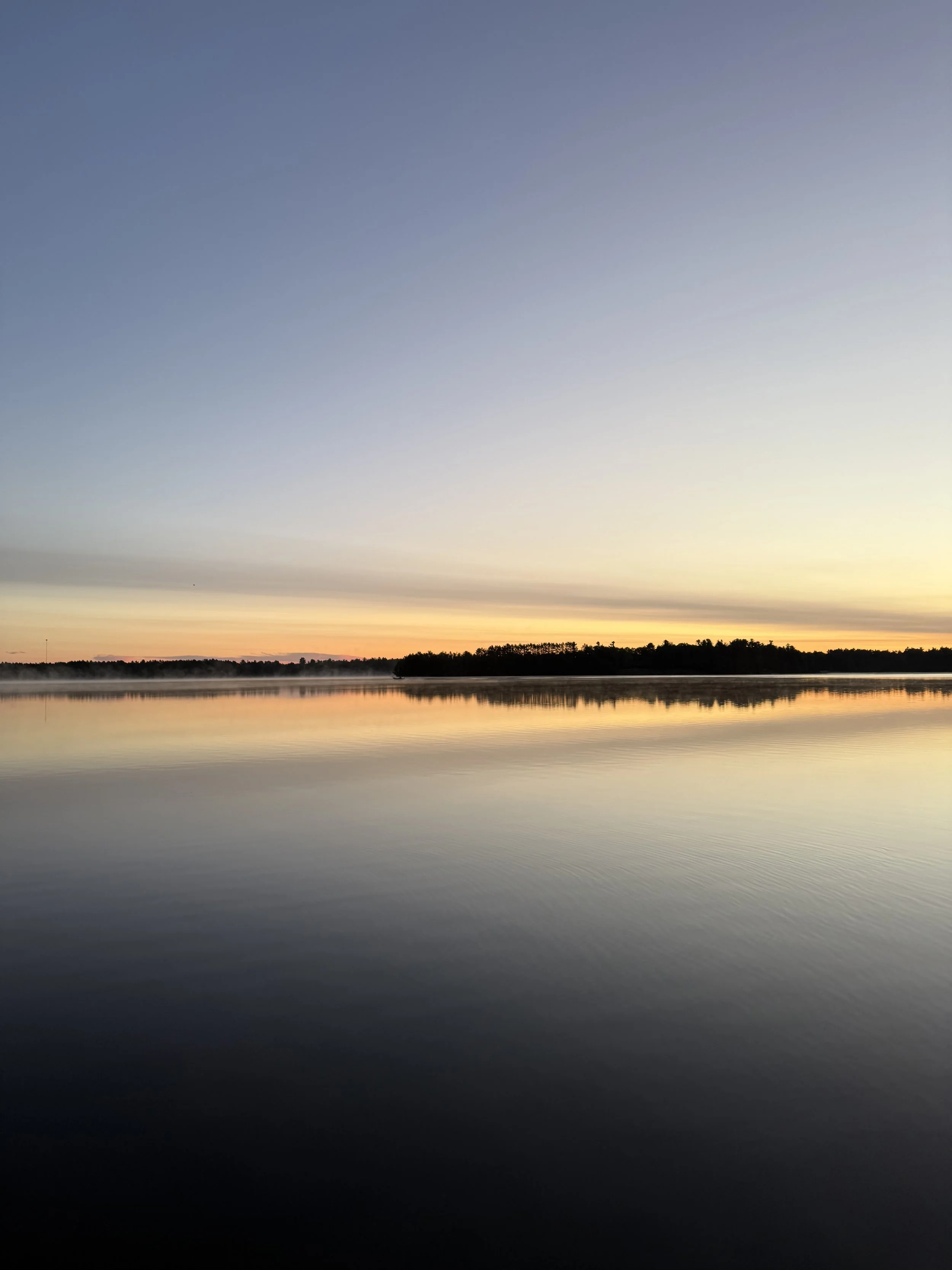 Serene lake at sunset with mist over the water, a tree-lined island, and reflections in the calm surface