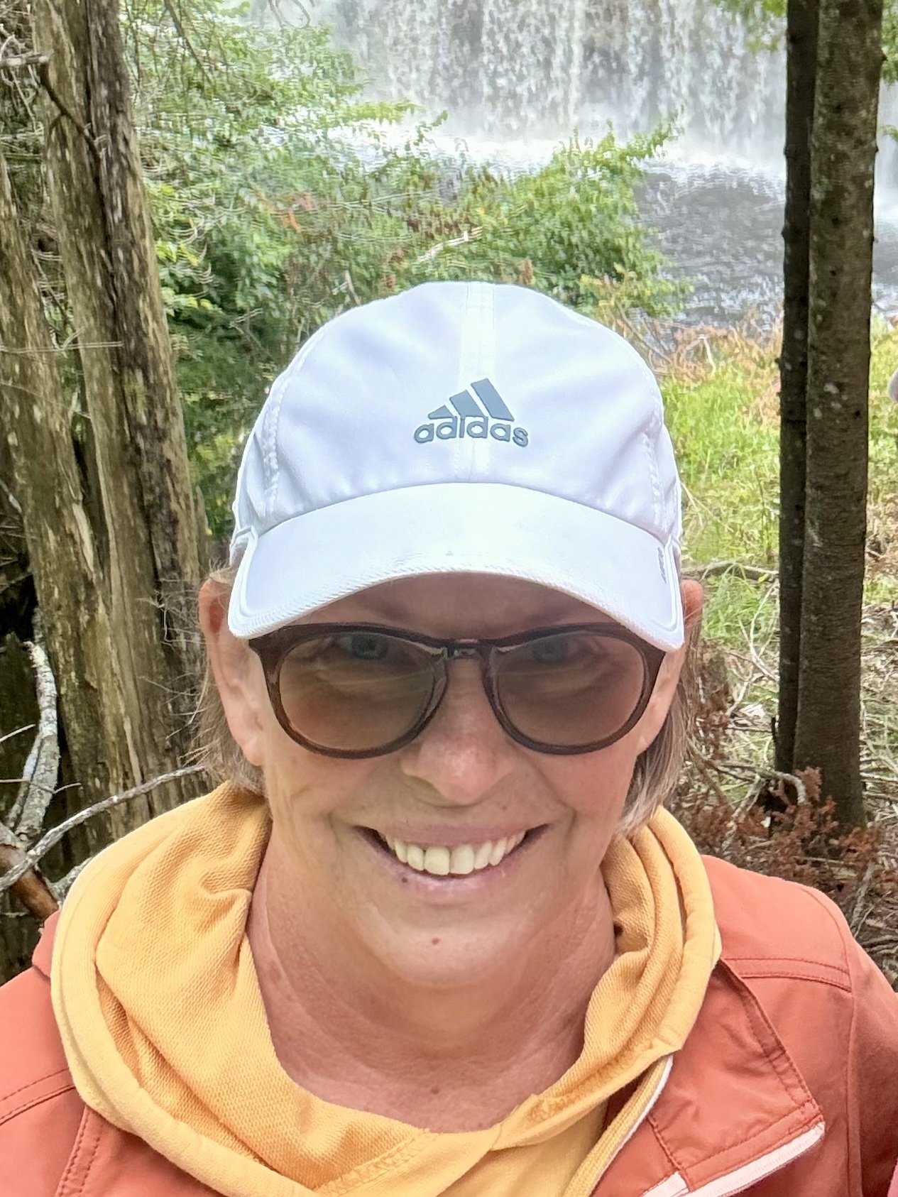 Smiling hiker wearing an Adidas cap and sunglasses in front of a forest waterfall