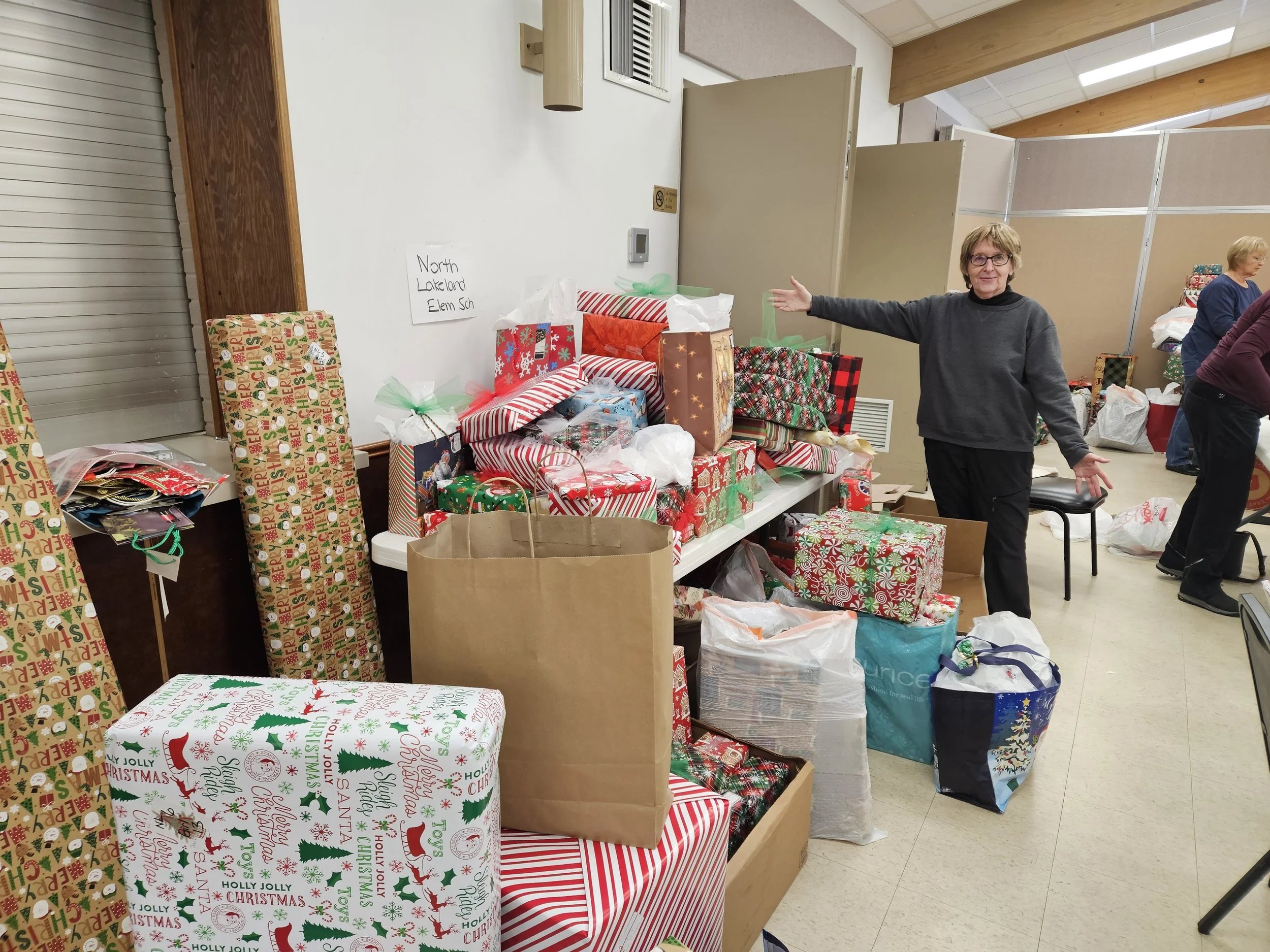 Volunteers organize piles of Christmas gifts and wrapped packages in a school room labeled North Lakeland Elementary School.