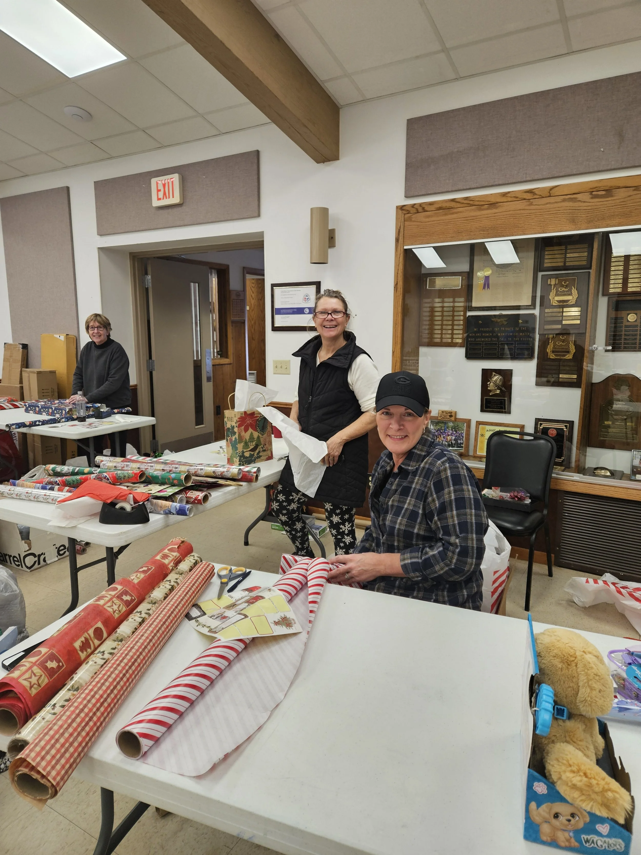Three women wrap Christmas presents at tables covered with colorful wrapping paper inside a community room.