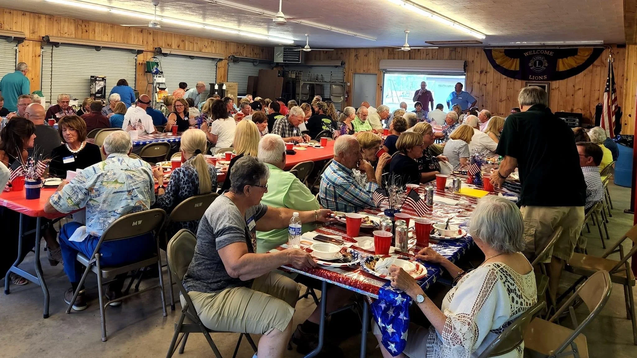 Crowded Lions Club gathering with people dining at American flag-themed tables in a community hall