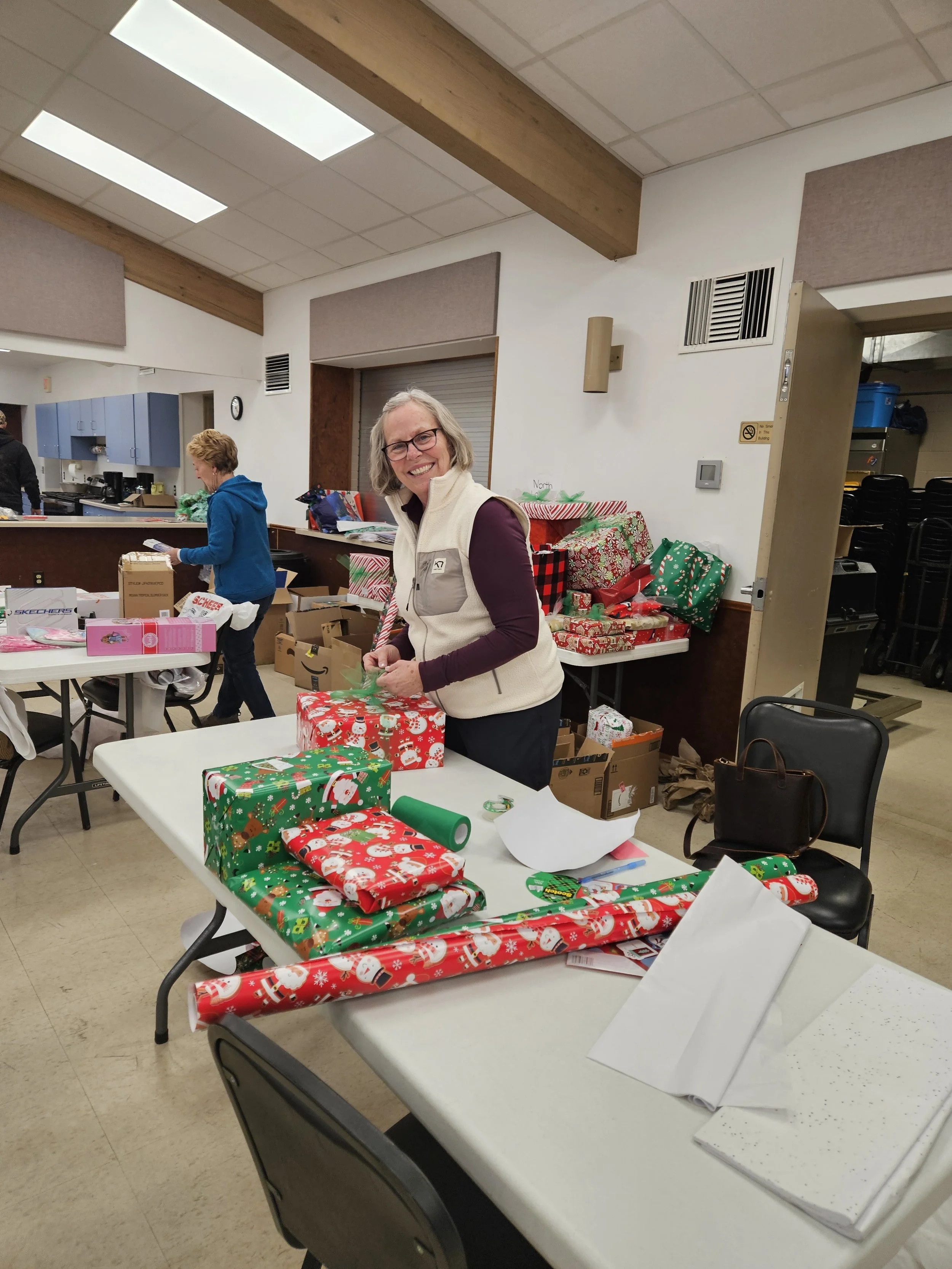 Volunteers wrap Christmas gifts at tables in a community room filled with presents and wrapping paper.