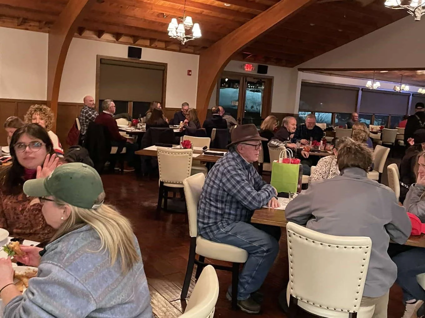 Crowded restaurant dining room with patrons seated at tables beneath a rustic wood-beam ceiling