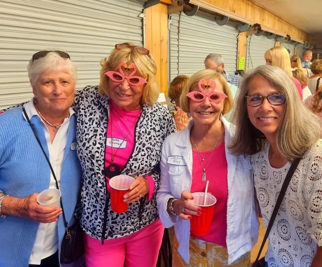 Four smiling women posing together at an outdoor gathering, two wearing pink novelty glasses and holding red cups.