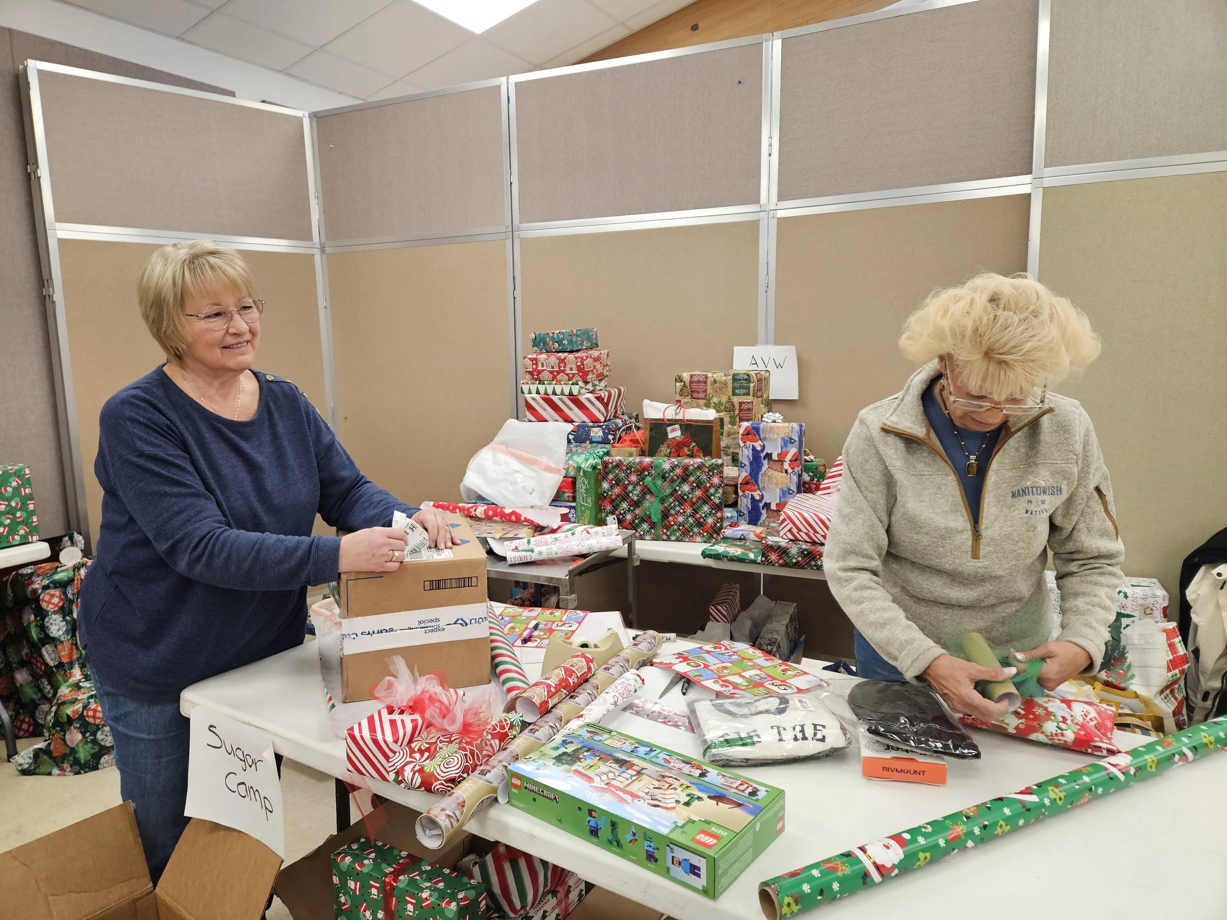 Two women wrap Christmas gifts at tables covered with presents, wrapping paper, and craft supplies in an office room.