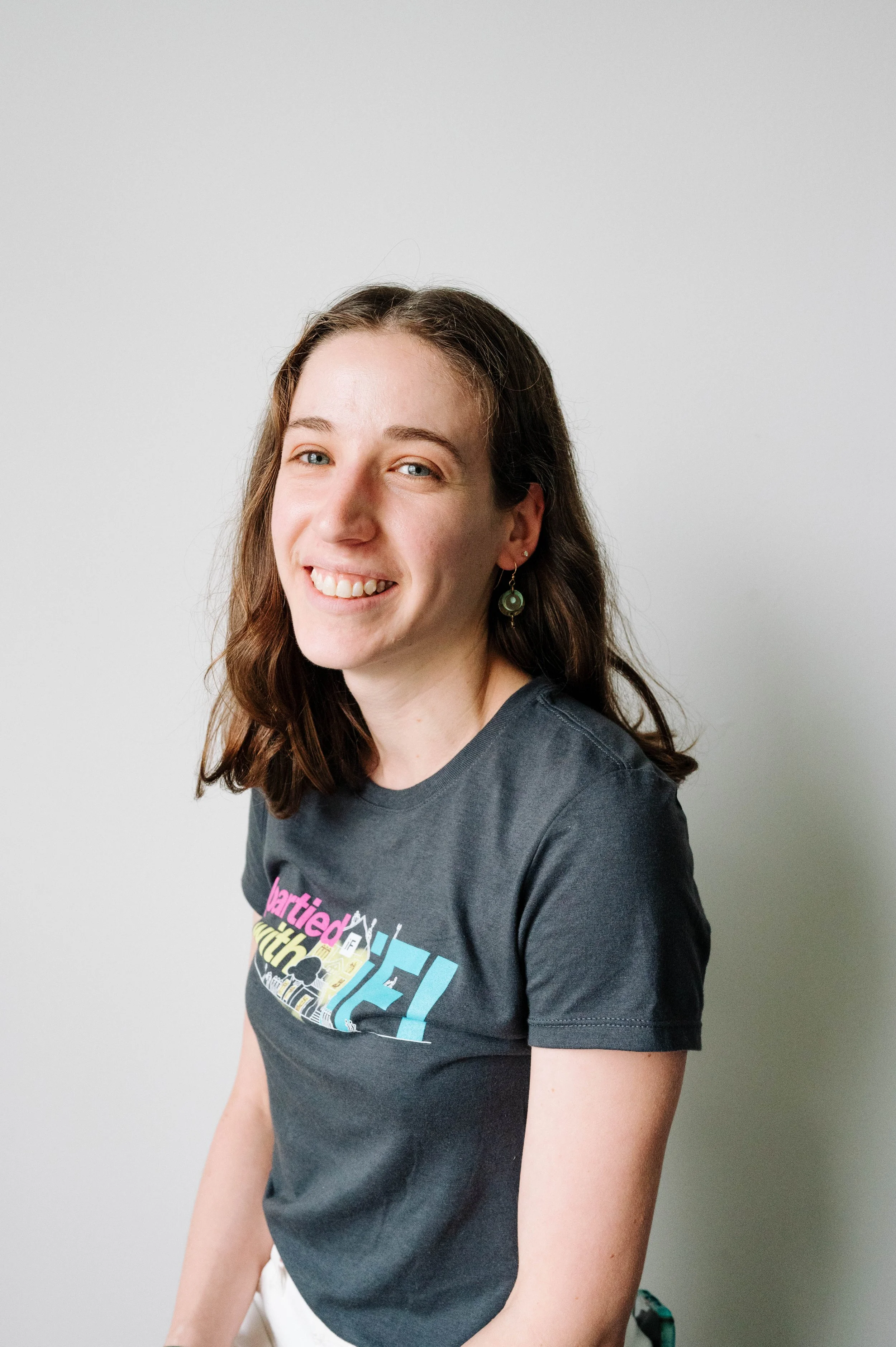 A woman with shoulder-length brown hair and earrings, smiling and wearing a dark graphic T-shirt, standing against a plain light gray background.