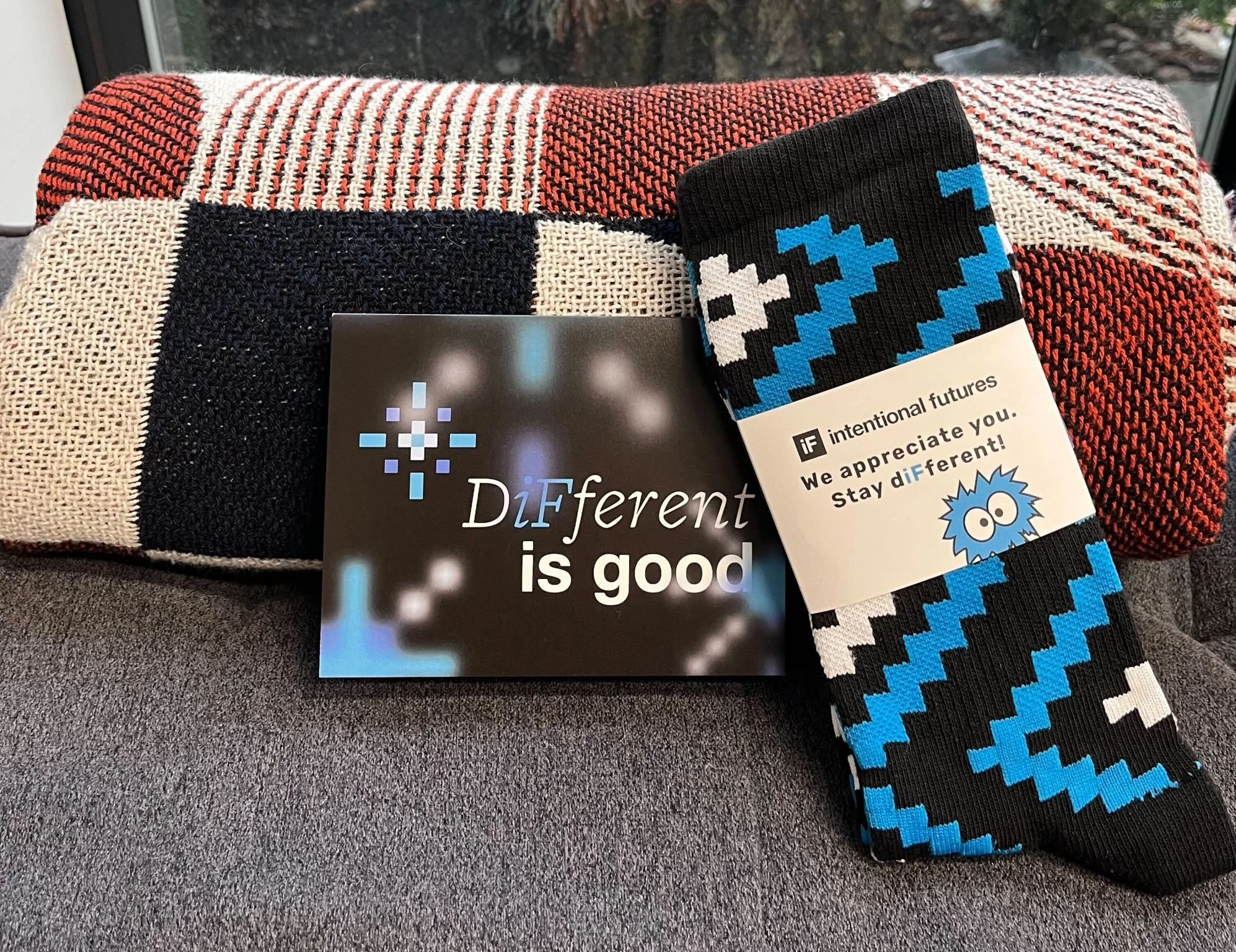 A colorful, patterned blanket with red, white, black, and cream colors drapes over a surface. A pair of black socks with blue, white, and black pixelated patterns is placed on top of the blanket. Part of a black booklet or flyer with the phrase "Different is good" is visible in front of the socks. The background shows a window with an outdoor scene.
