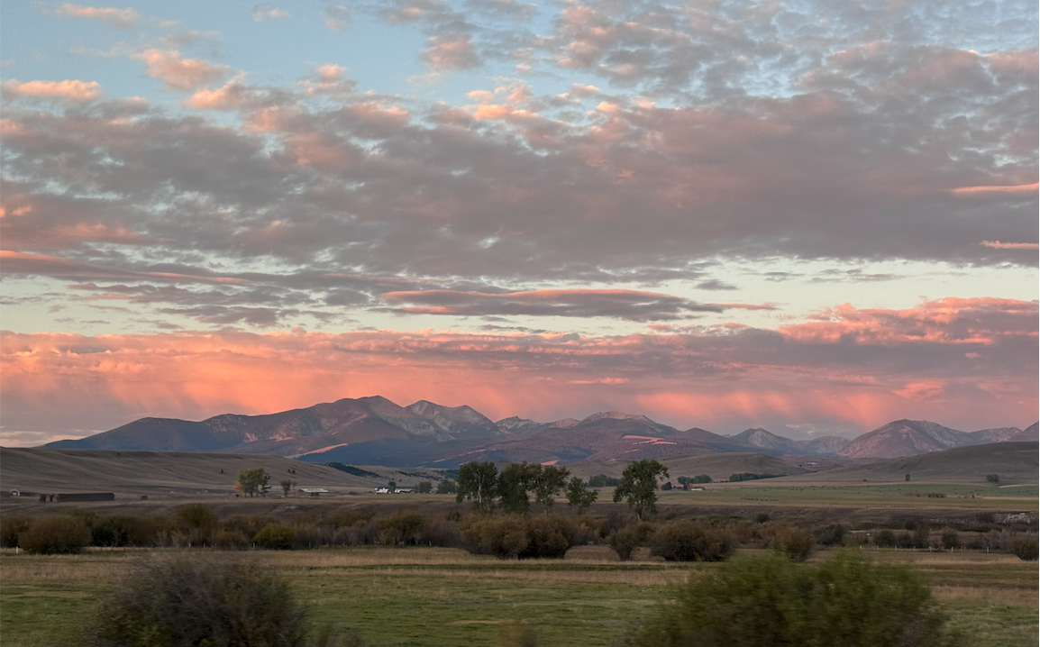 Scenic landscape with mountains at sunset, pink and orange clouds, green fields, and scattered trees.