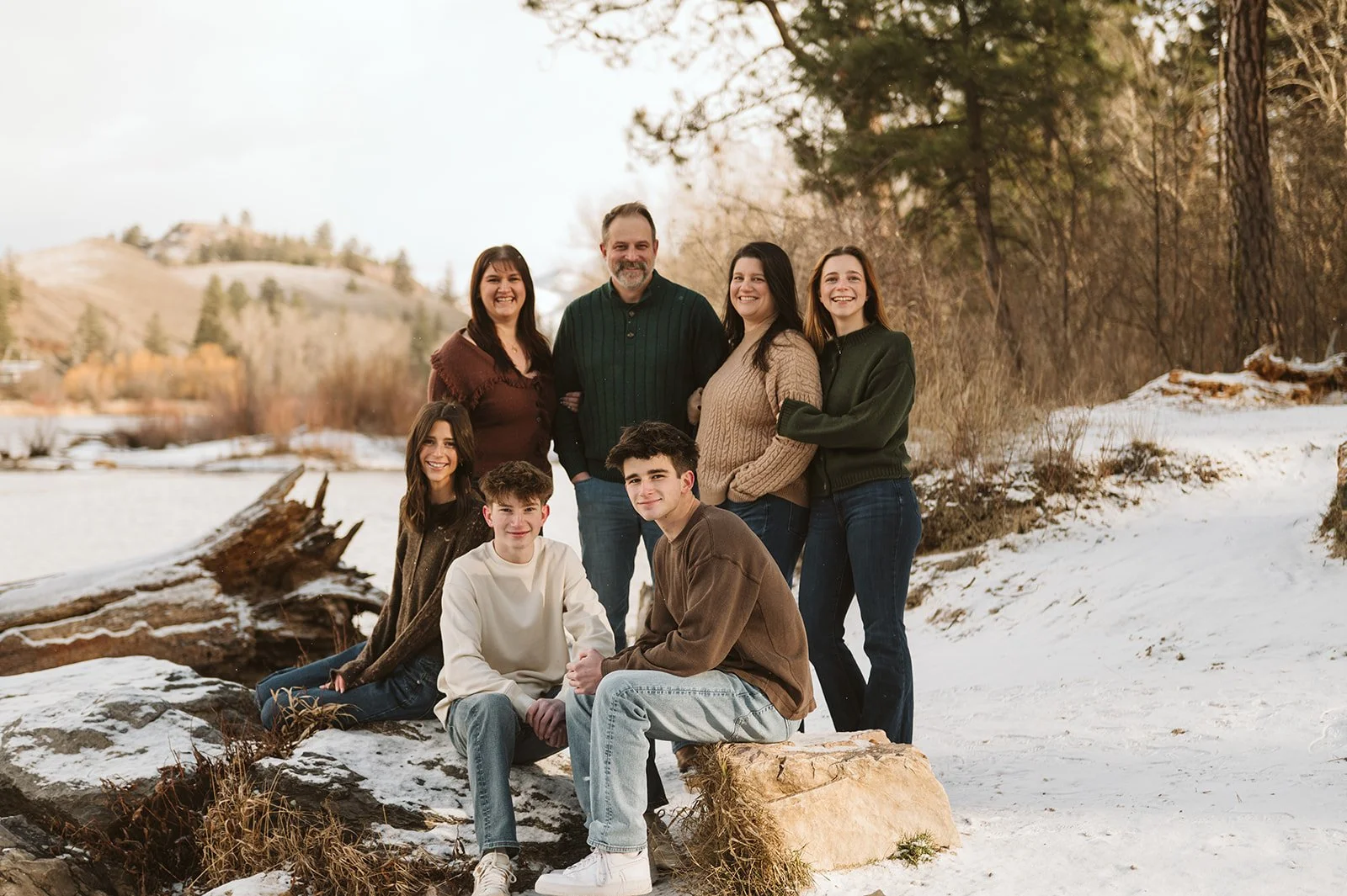 A family of seven poses outdoors in a snowy landscape with a river, trees, and hills in the background. The family members are smiling and dressed in winter clothing.