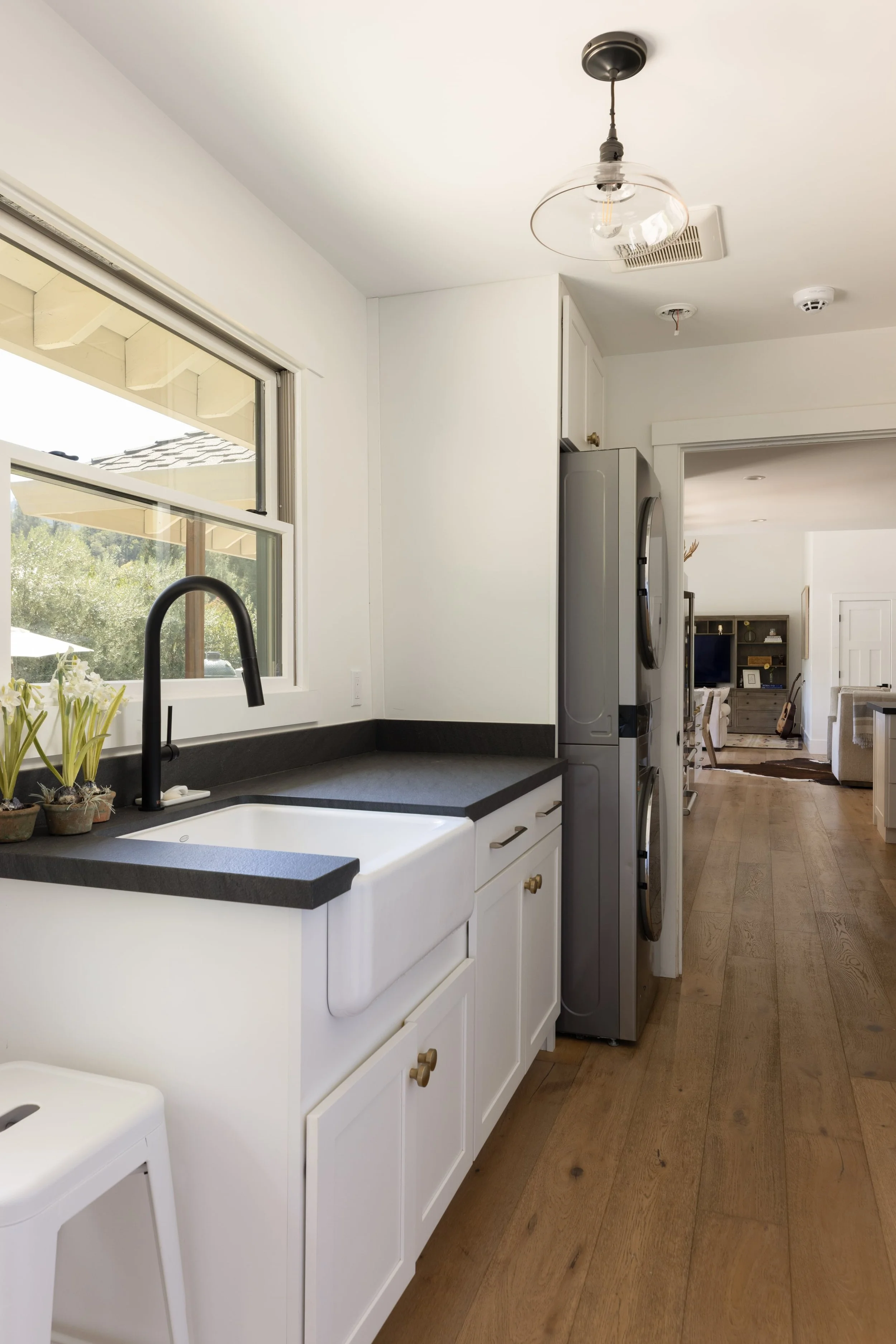 Kitchen with white cabinets, black countertop, farmhouse sink, black faucet, window showing outdoor patio, and wooden flooring.