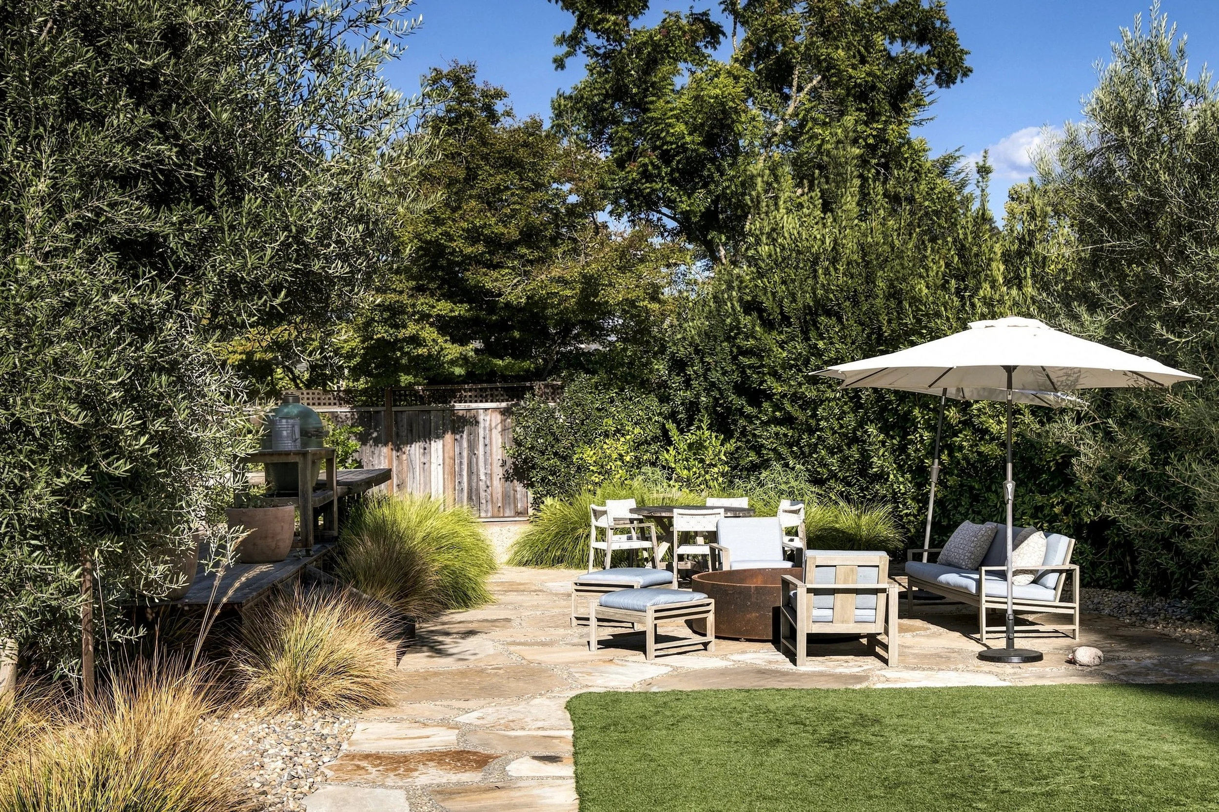 A backyard patio with outdoor furniture including chairs, a table, and an umbrella, surrounded by lush green trees and plants under a blue sky.