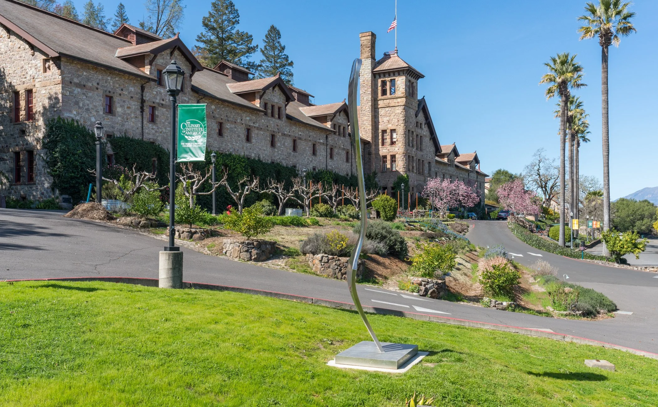 The image shows a large, historic stone building with a tower, surrounded by palm trees and flowering trees. There is a banner indicating the culinary college, and a winding driveway leading up to the building with landscaped greenery and a street la