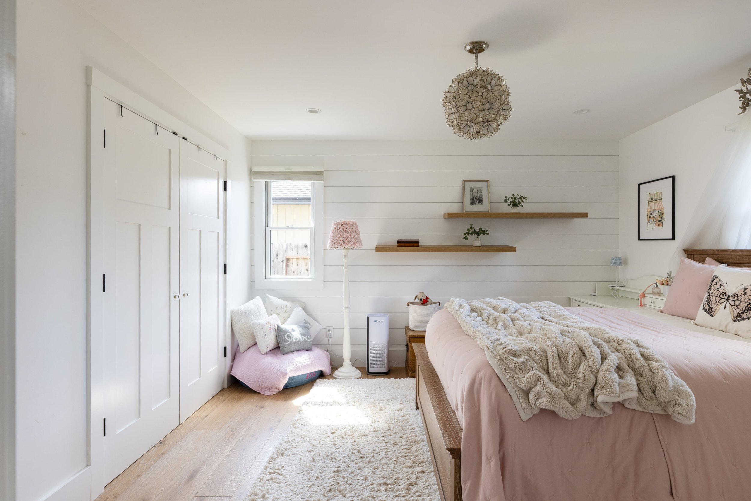 A bright bedroom with white walls and a large bed with pink bedding and a fluffy beige throw. There are two wooden floating shelves on the wall with framed pictures and small plants. A window lets in natural light, illuminating a pink cushion, stuffe