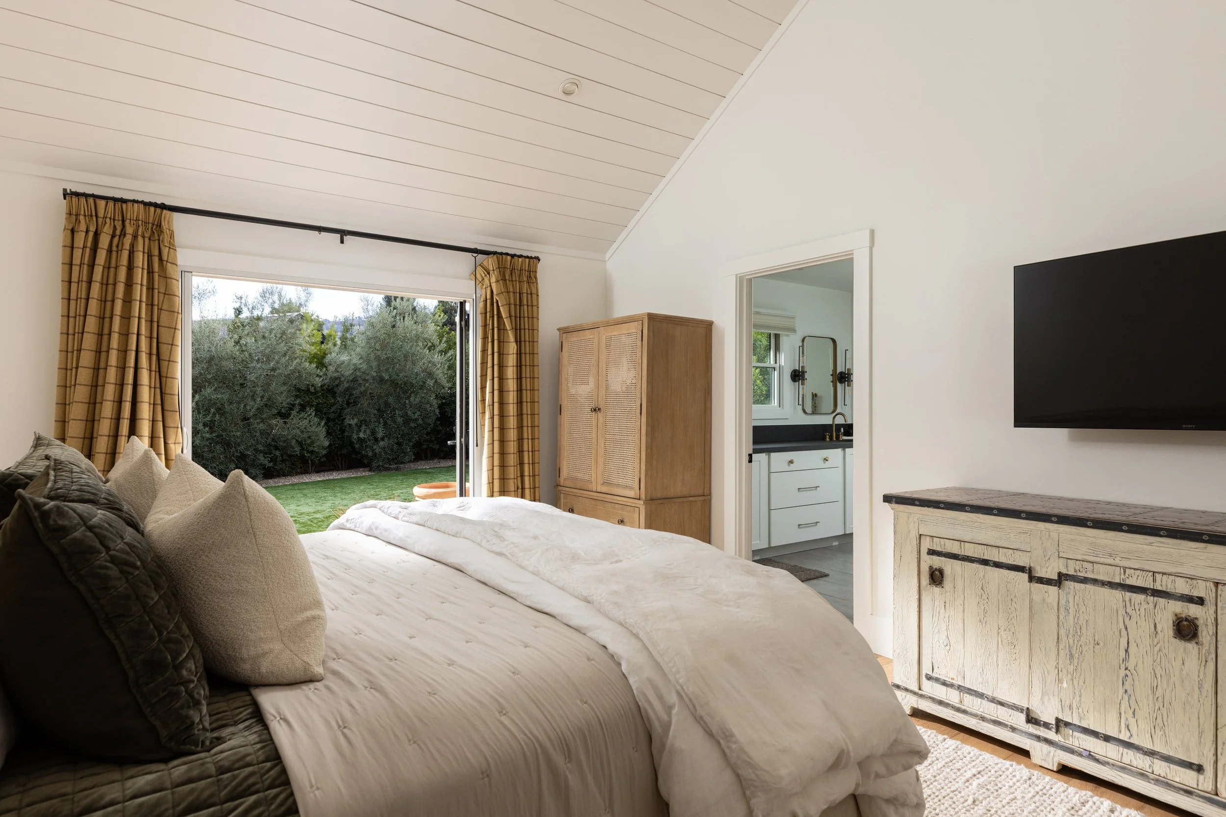A bedroom with a large bed covered in white bedding and multiple pillows, a window with beige plaid curtains open to a view of trees, and a wooden cabinet. There is a doorway leading to a bathroom with a window, a mirror, and a sink. A flat-screen TV