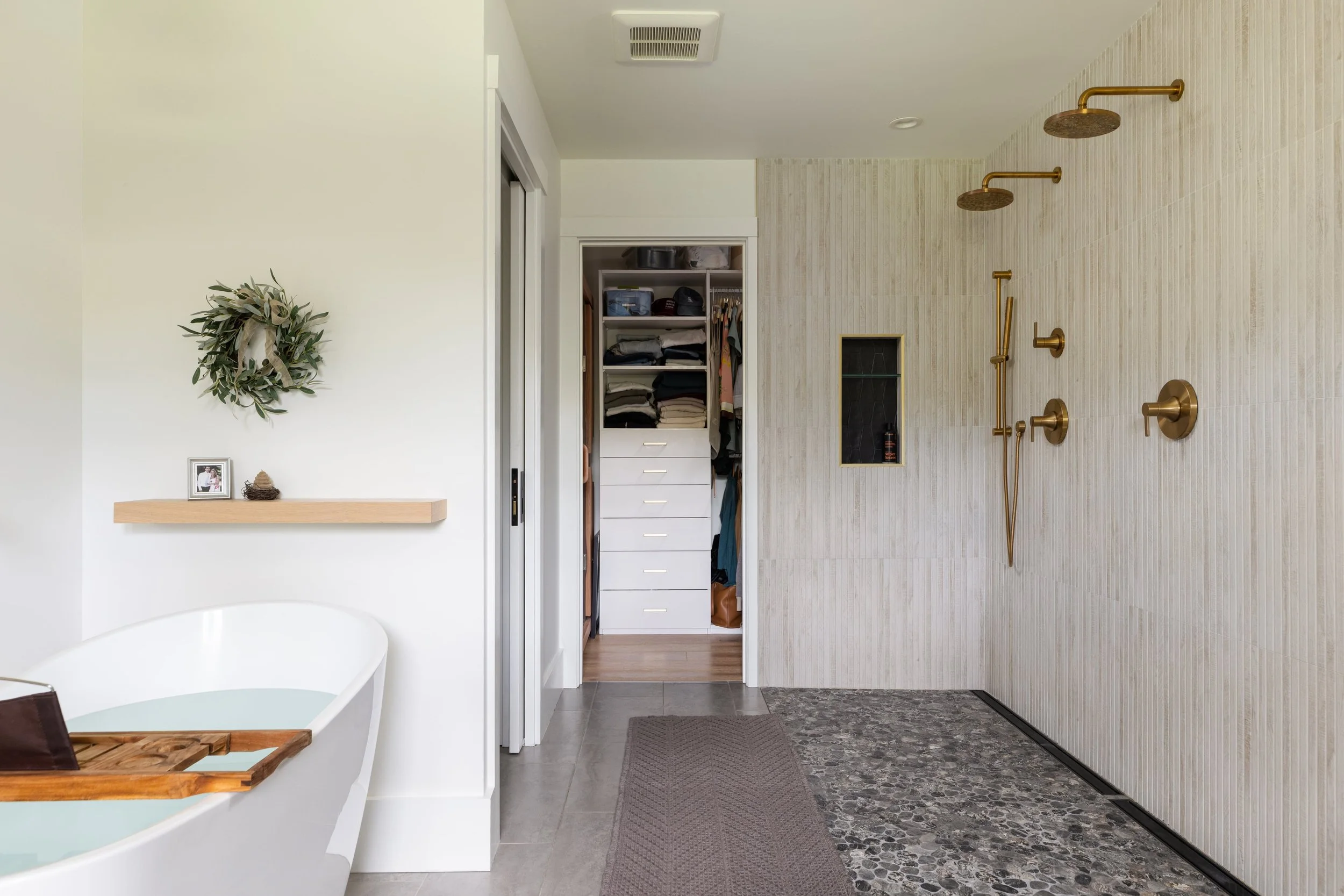 Modern bathroom with a white bathtub, a floating wooden shelf with a framed photo, a greenery wreath, an open closet with folded clothes and hanging garments, a textured tile shower wall with three gold showerheads and controls, and a small dark nich