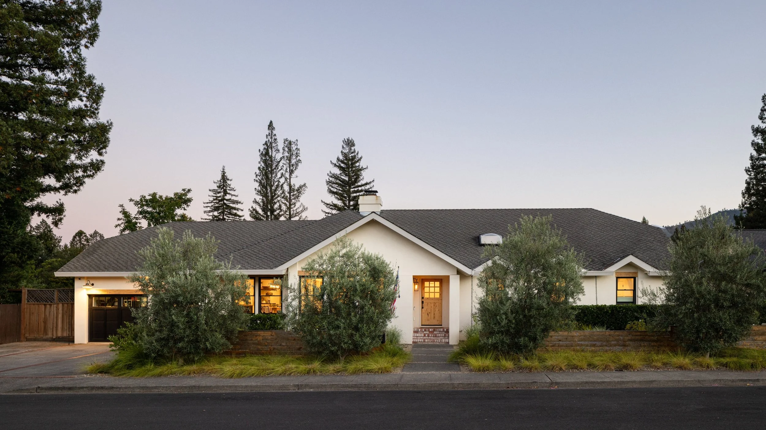 Modern suburban house with a dark gray roof, white exterior walls, and illuminated interior lights, surrounded by trees and a sidewalk in the evening.