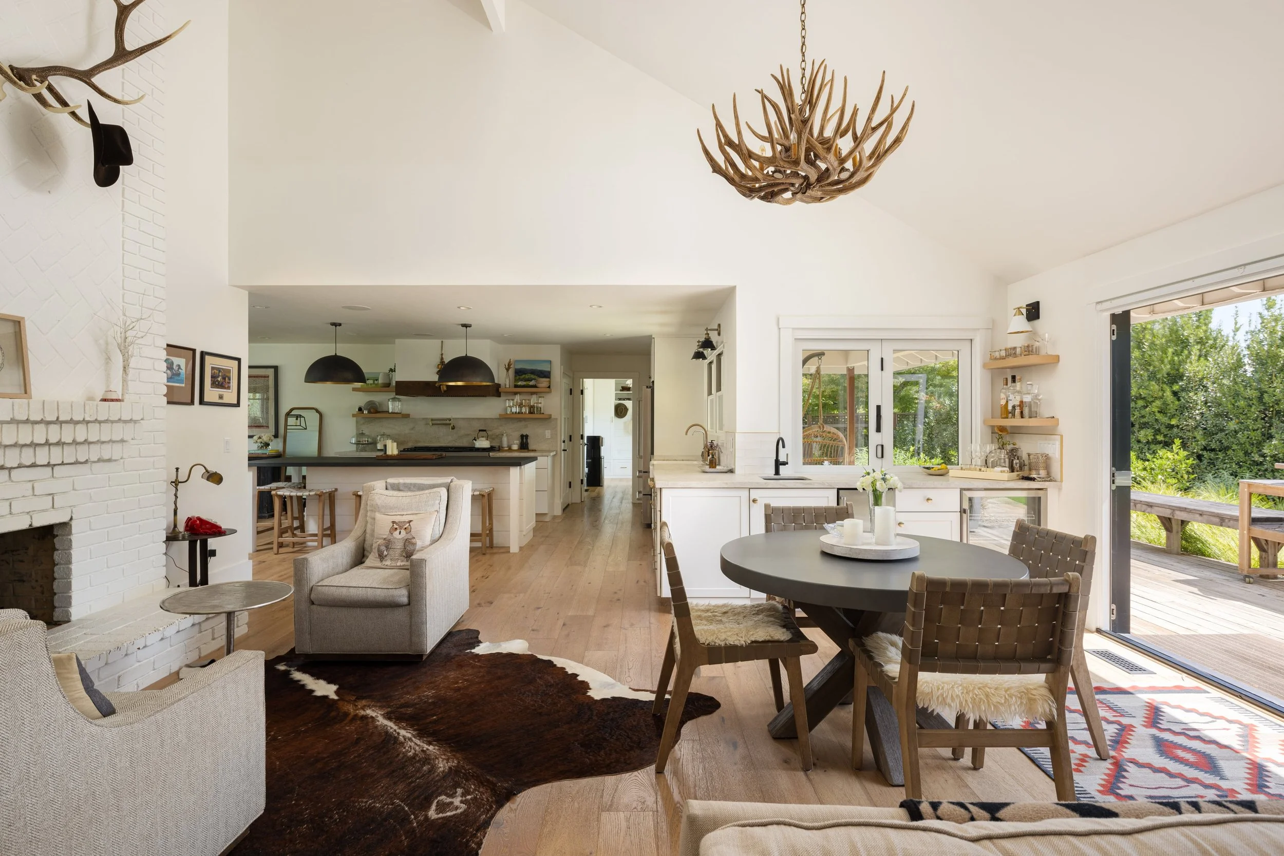 Bright open-concept kitchen and living room with white walls, wooden floors, and large windows and sliding doors opening to a deck with greenery outside. Features include a white fireplace, black pendant lights, a round dining table with wicker chair