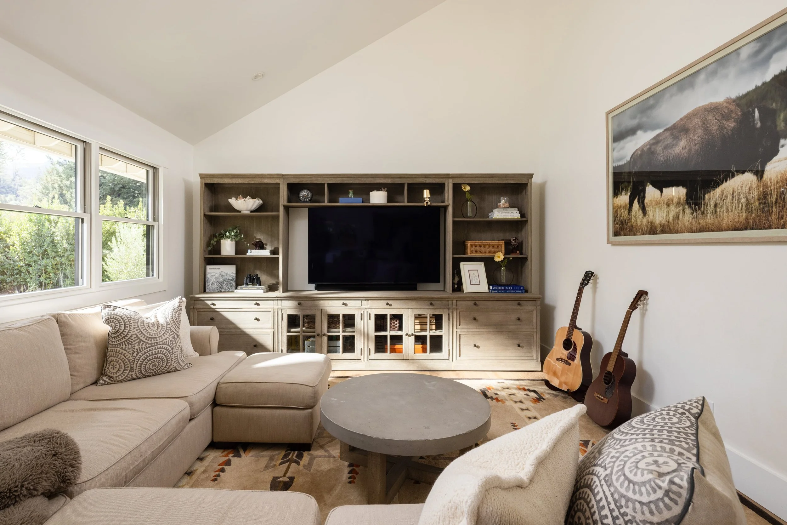 Living room with beige couch, patterned pillows, coffee table, large windows with greenery outside, wall-mounted wooden entertainment center with TV, decorative items, framed photo, guitar, and large framed mountain landscape artwork on the white wal